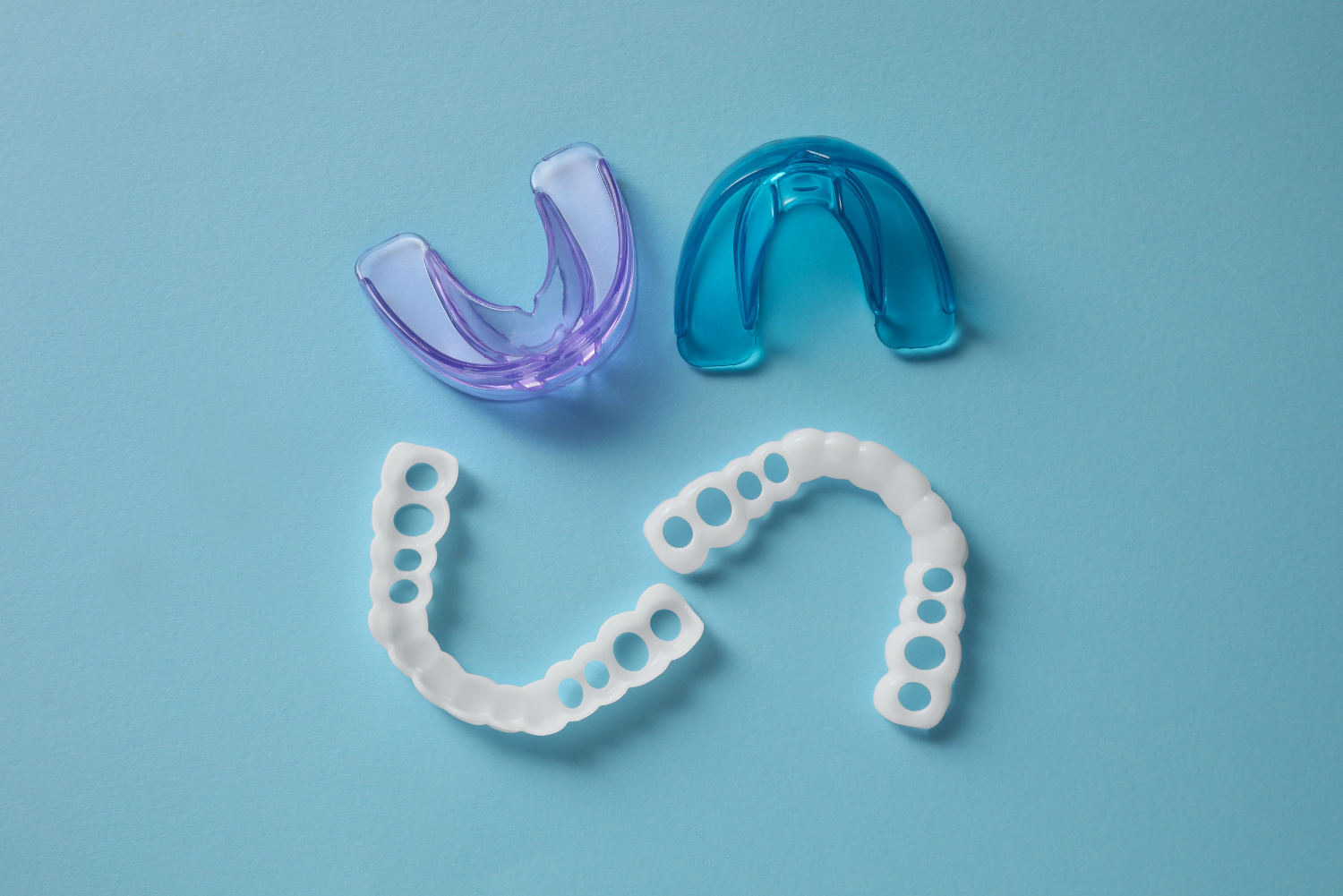 Two clear aligners, one purple and one blue, beside two white dental molds on a light blue surface