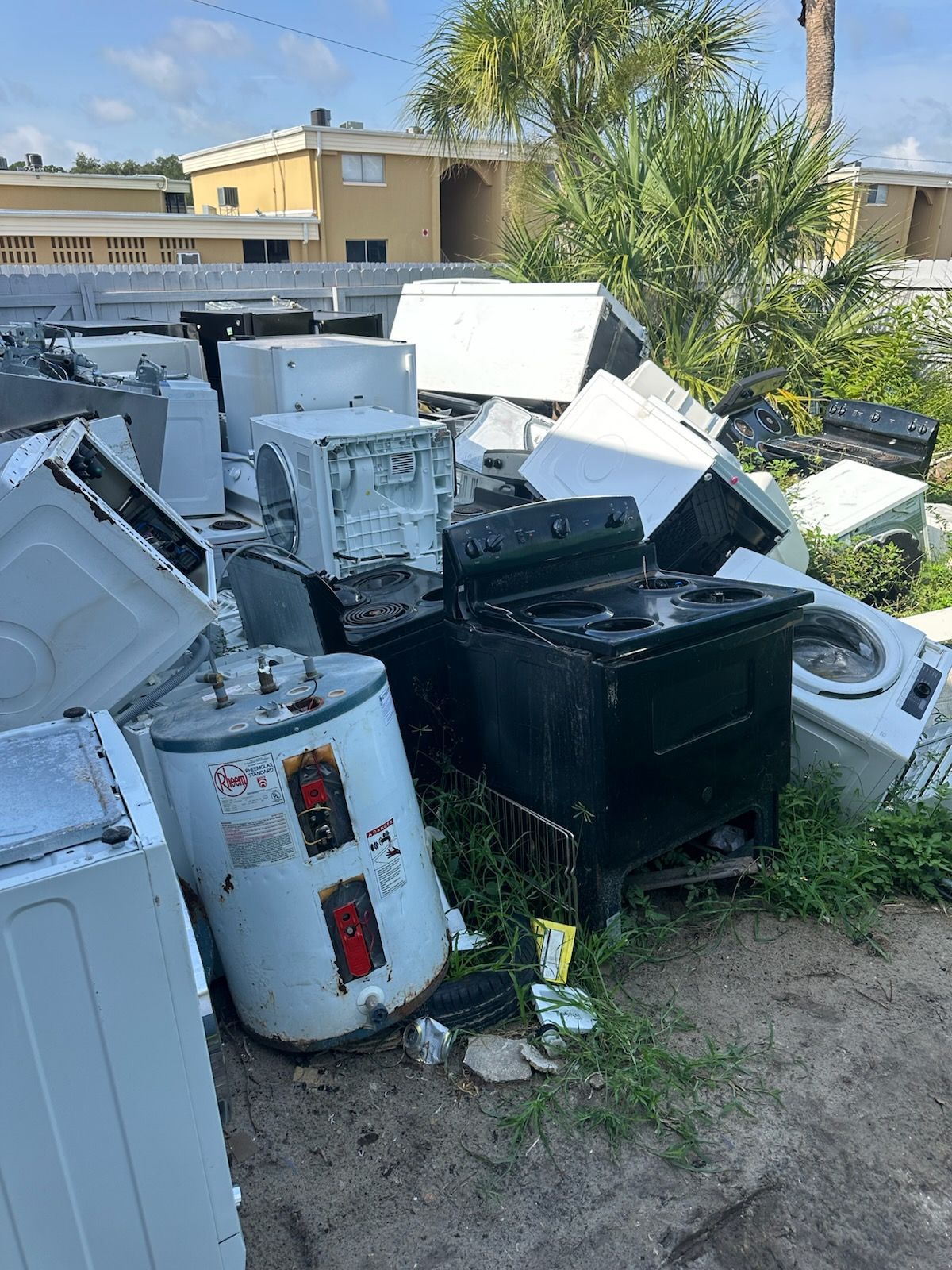Pile of discarded appliances and scrap in a yard, with white and black machines stacked near a house.