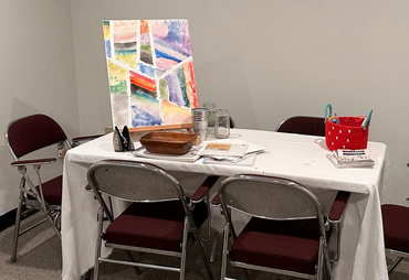 The picture shows a table with tablecloth set for painting activities with a completed watercolor canvas on an easel and six folding chairs.