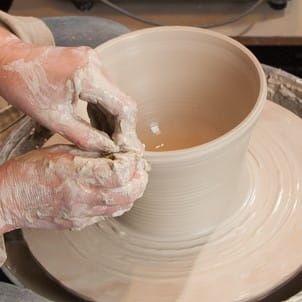 Handen vormen een kleipot op een pottenbakkerswiel, met natte klei en draaiende slib rond de basis.