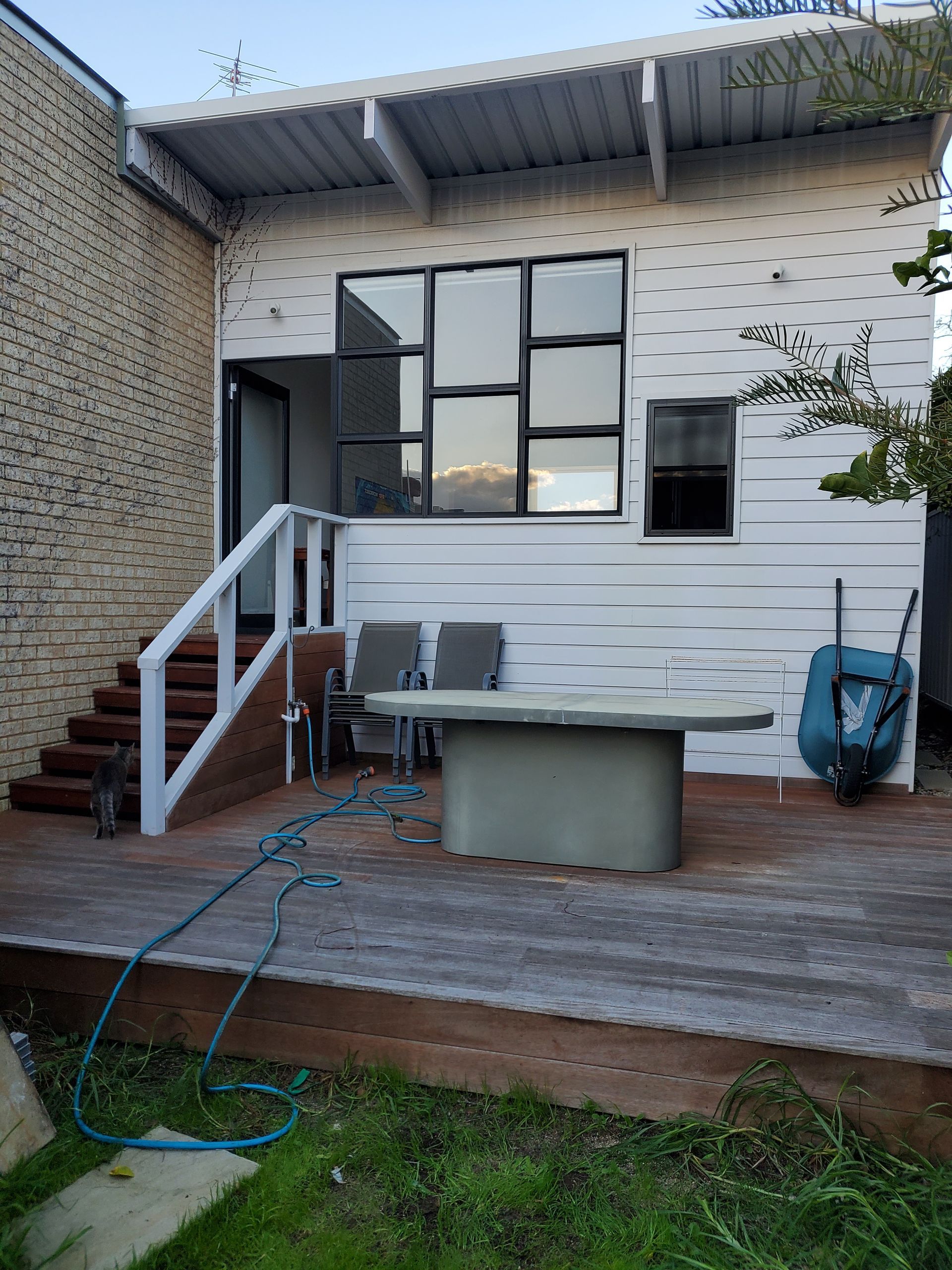 Wooden deck with stairs leading to a white building. A water hose, chairs, and wheelbarrow are visible.