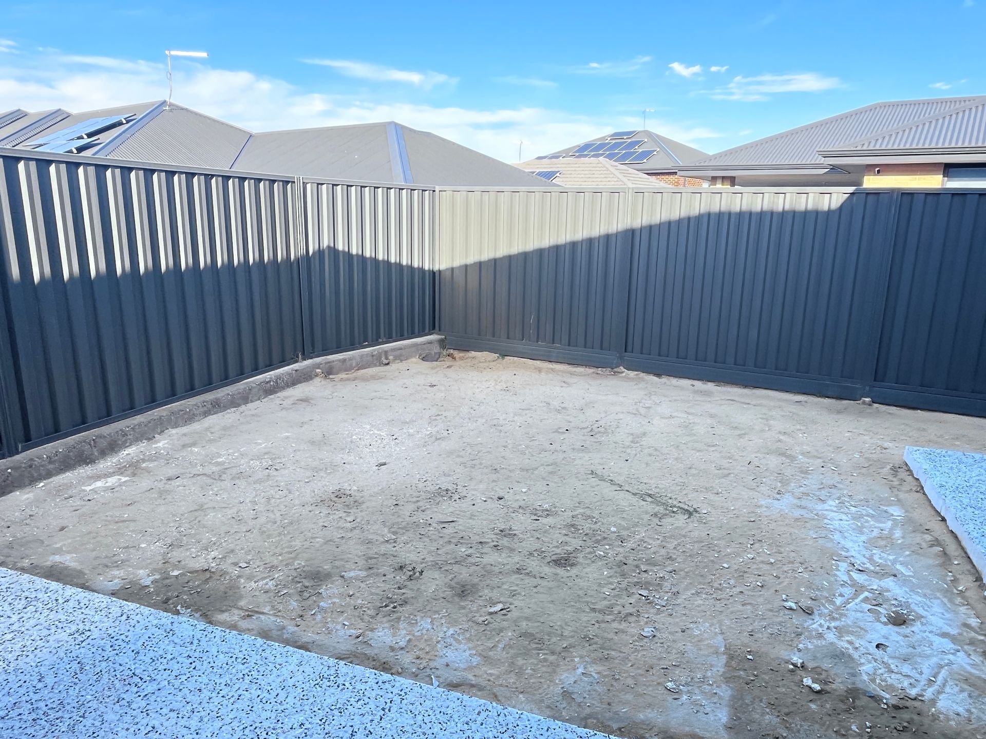 Empty backyard with a concrete base, enclosed by a grey fence, under a blue sky.