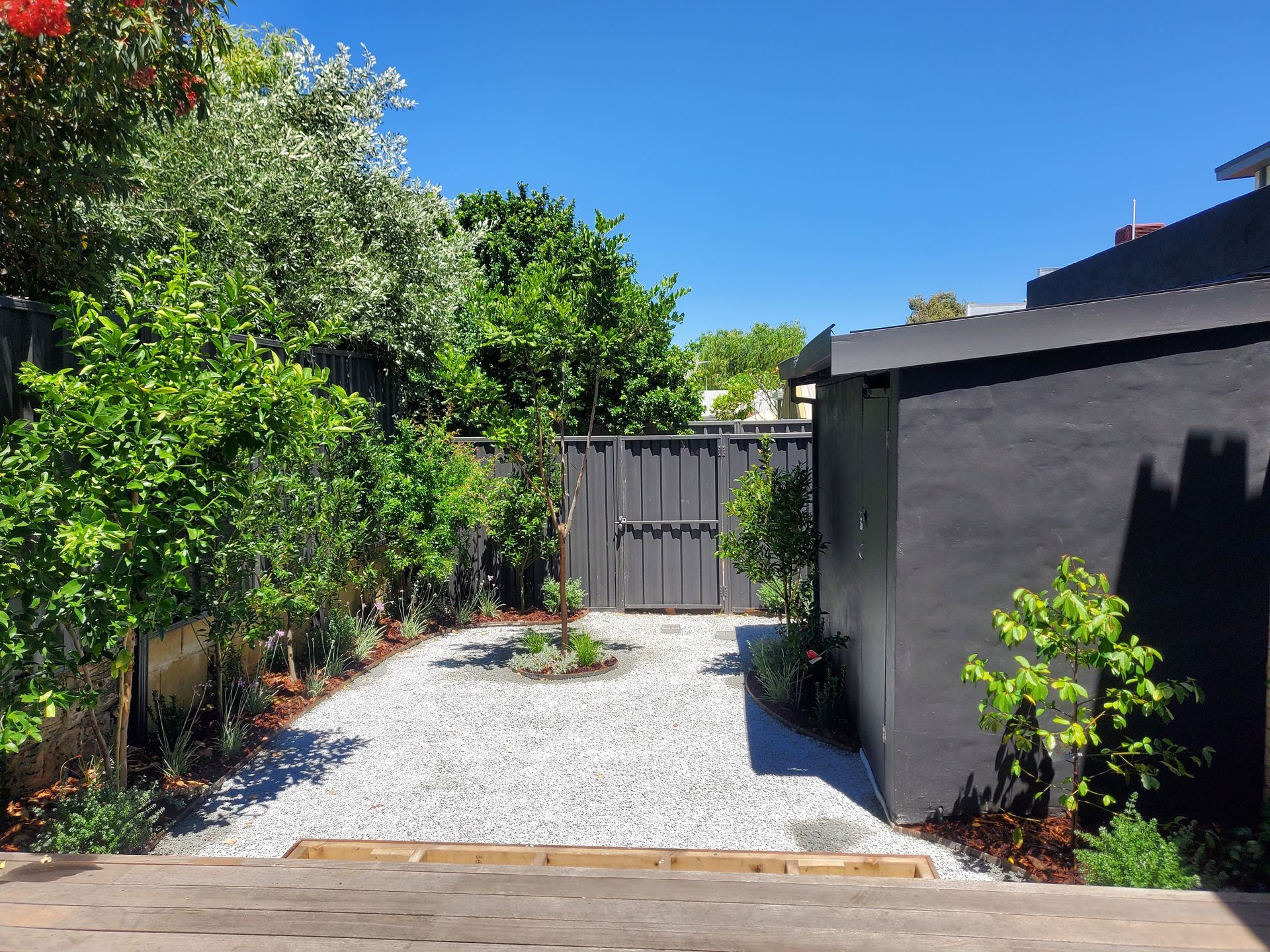 Gravel pathway leading to a dark gray fence, lined with greenery and a black building on the right. Blue sky.