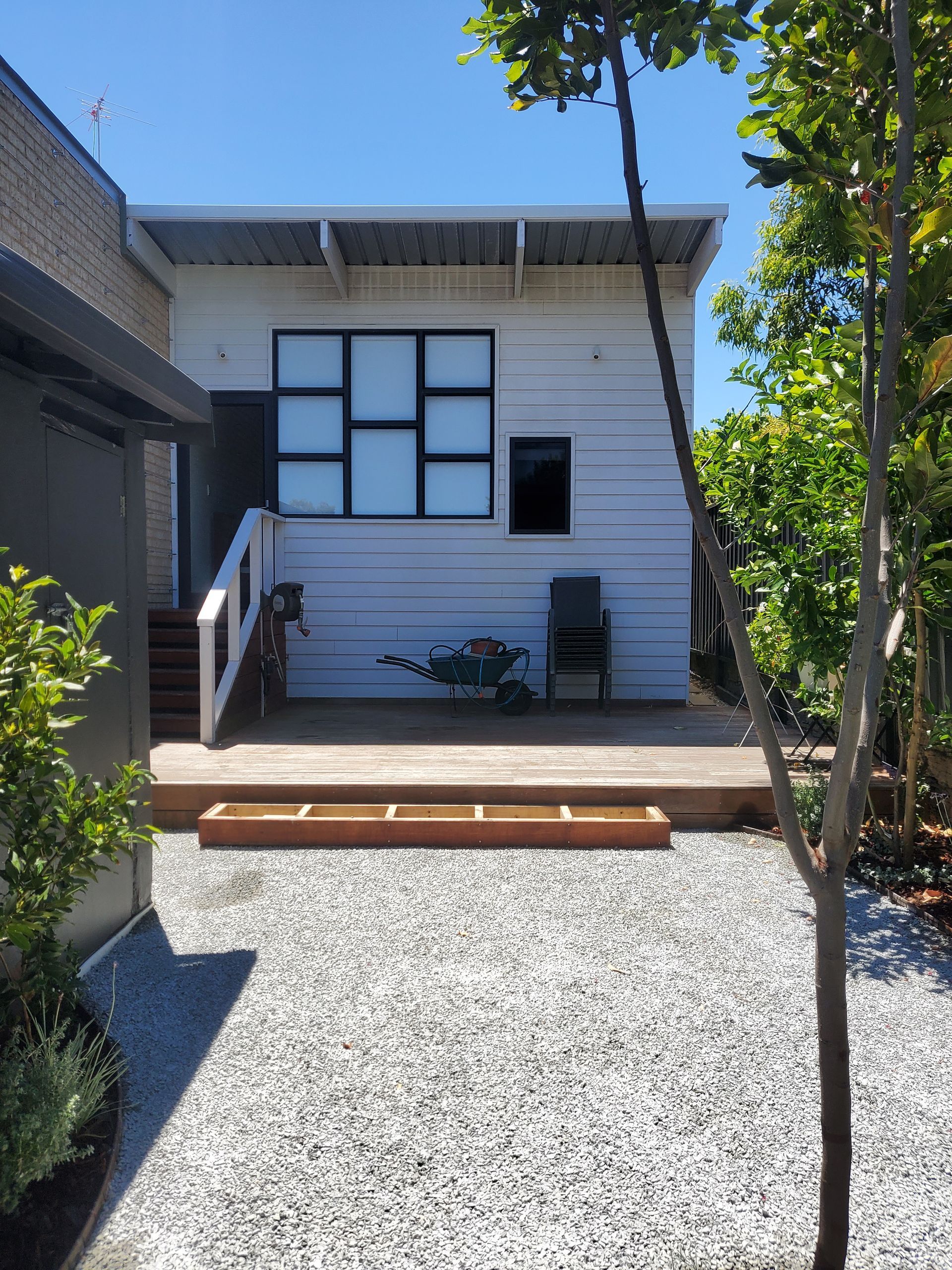 A backyard scene with a white building, wooden deck, gravel, and trees under a sunny sky.