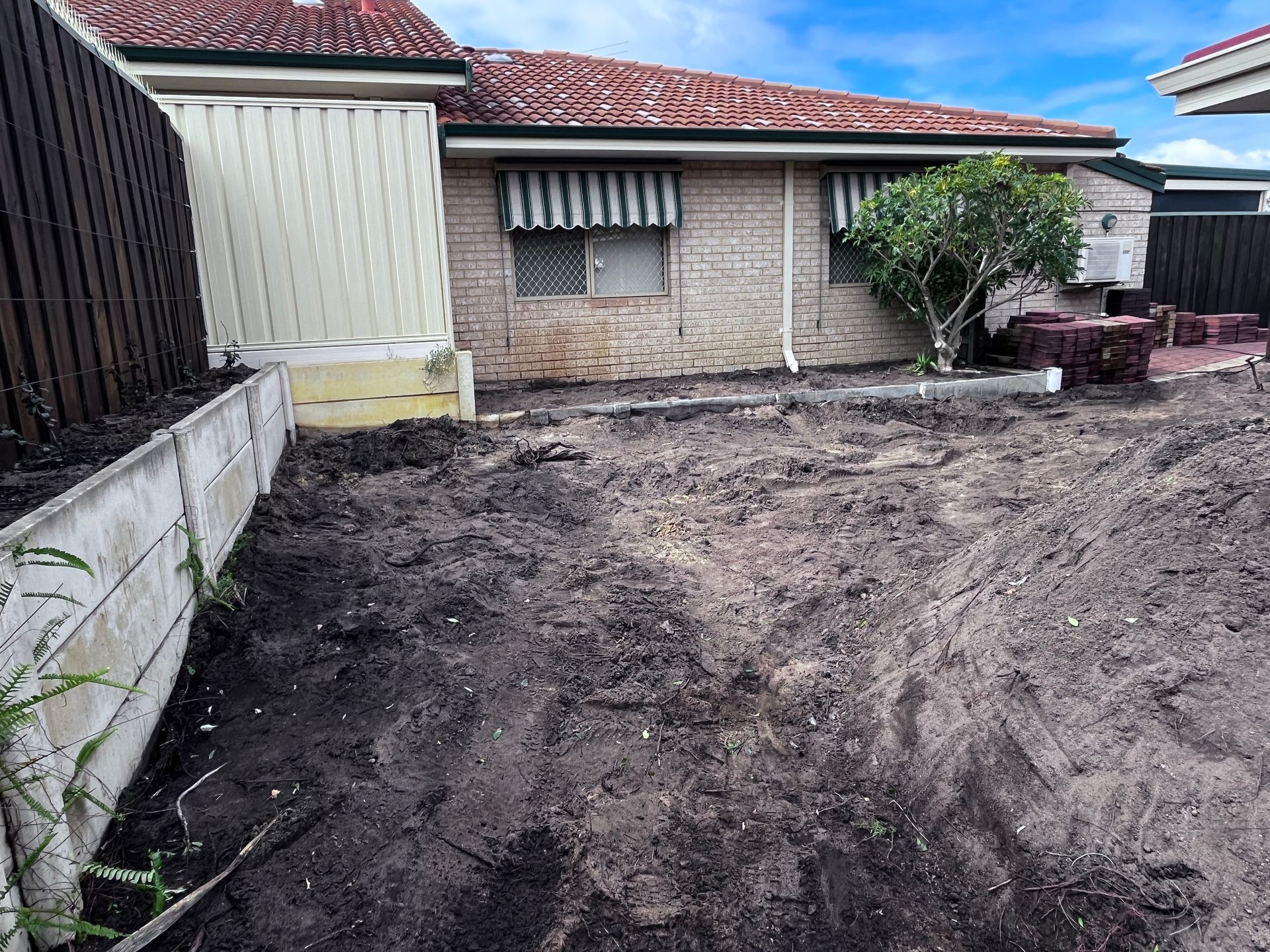 Backyard with exposed soil, brick house with awnings, and retaining wall.