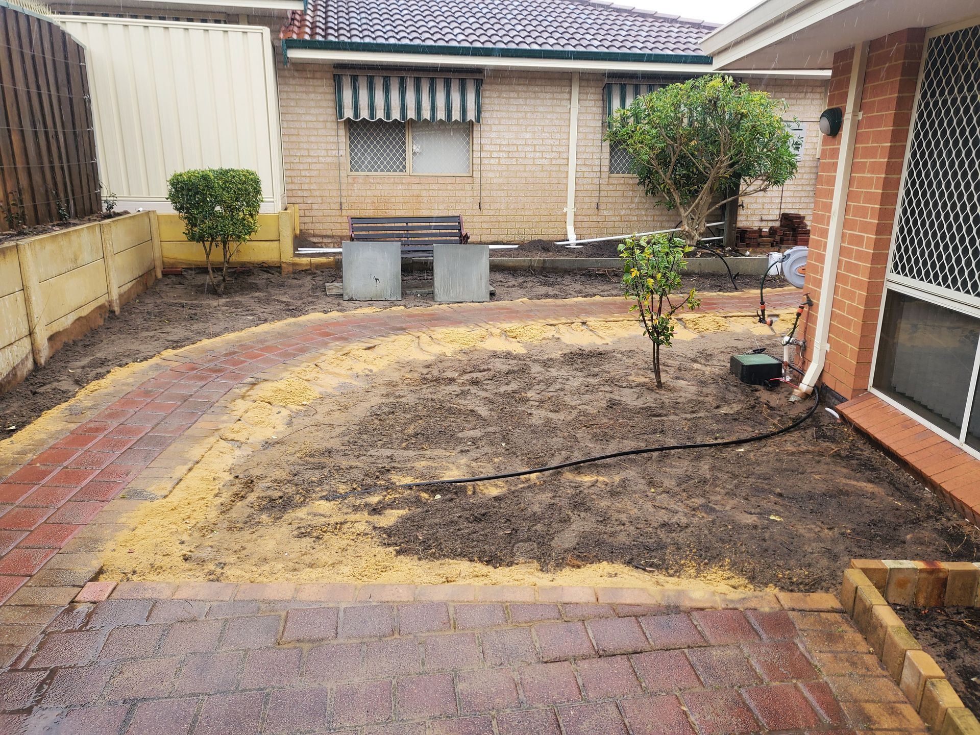 Brick pathway winds through a backyard garden with freshly turned soil, small plants, and a brick house.