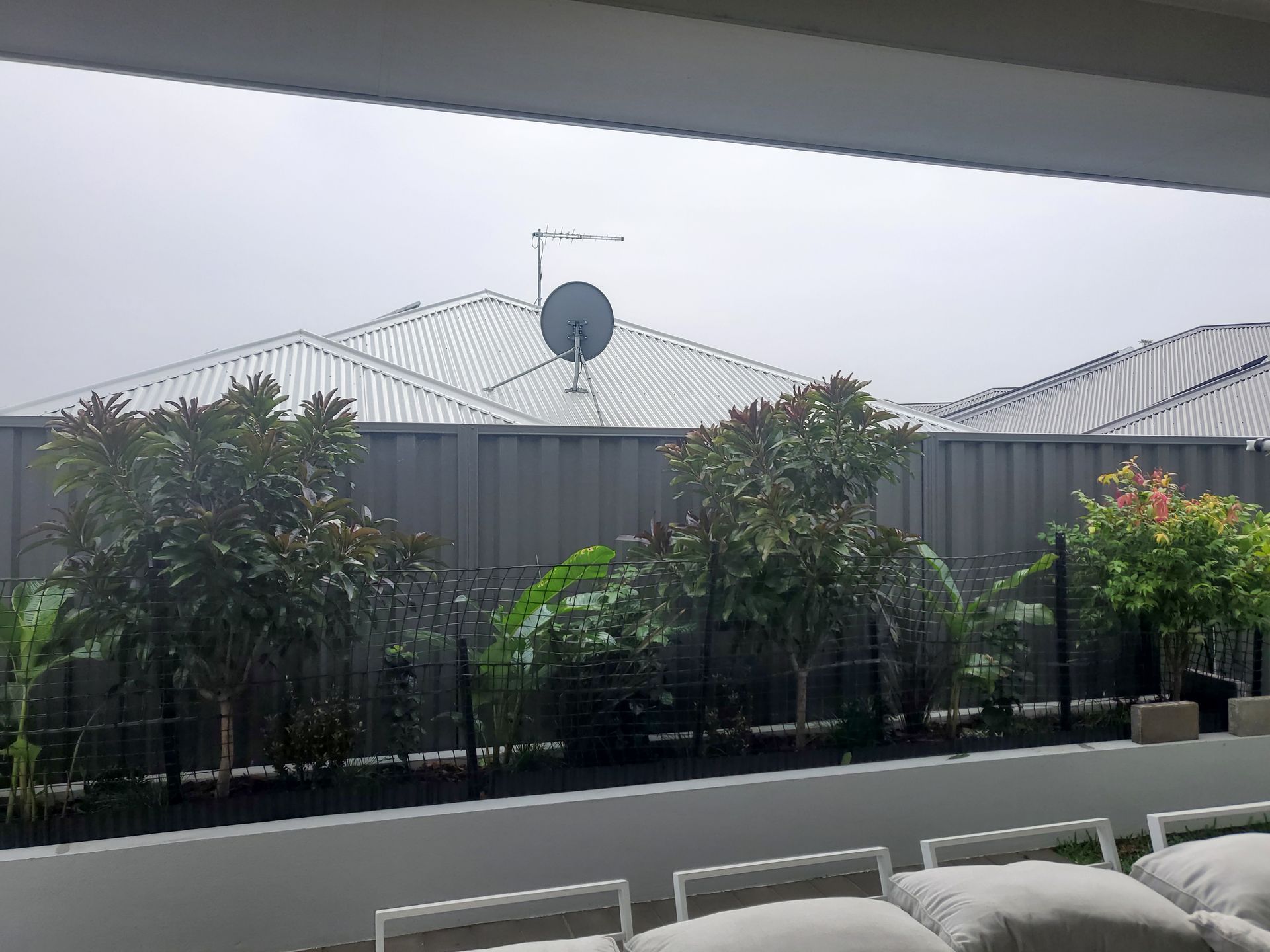 View from a patio of a backyard with trees, a fence, and a roof with a satellite dish. Cloudy sky.