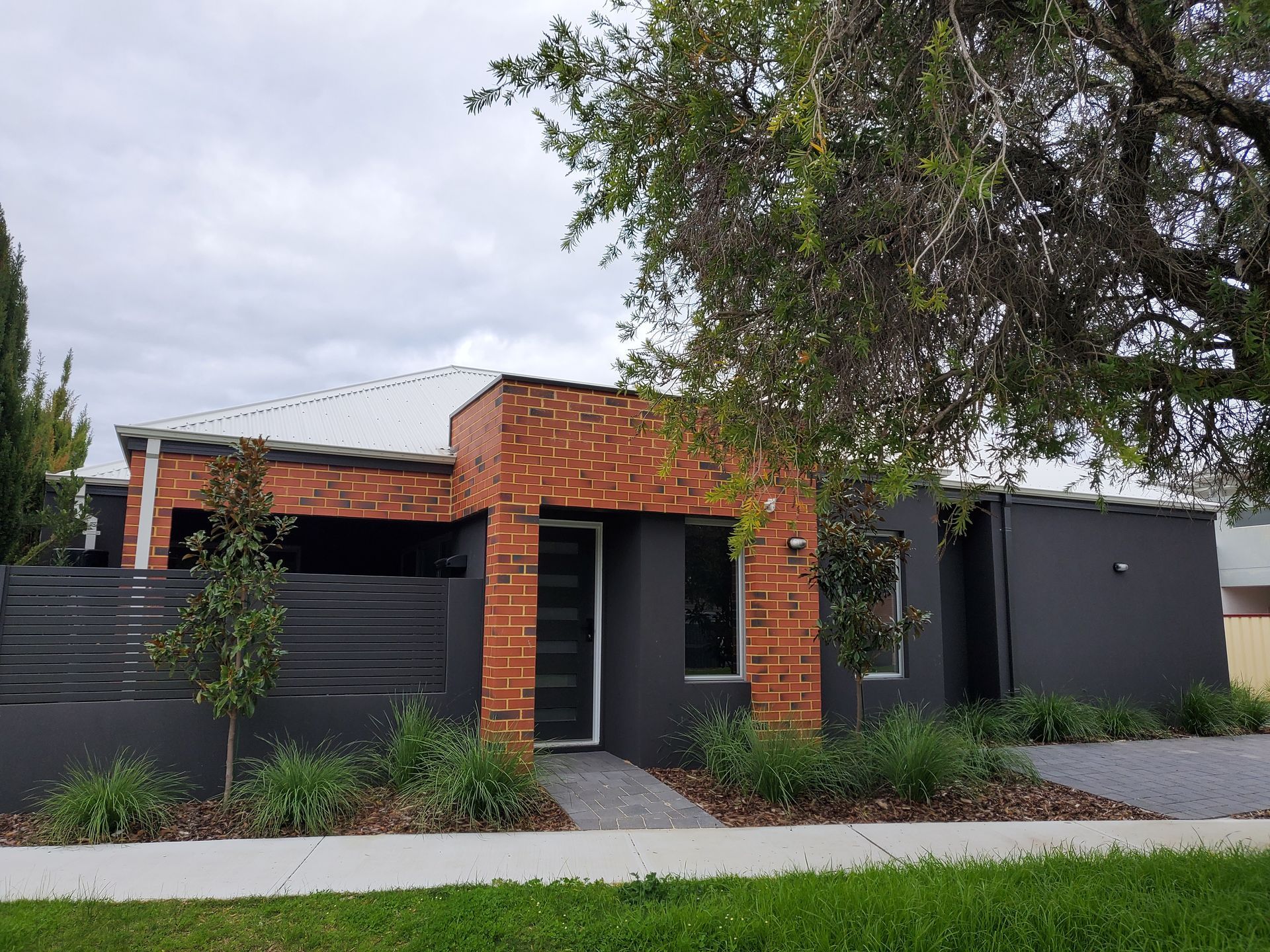 Modern house with brick accent, dark gray walls, and landscaping under a cloudy sky.
