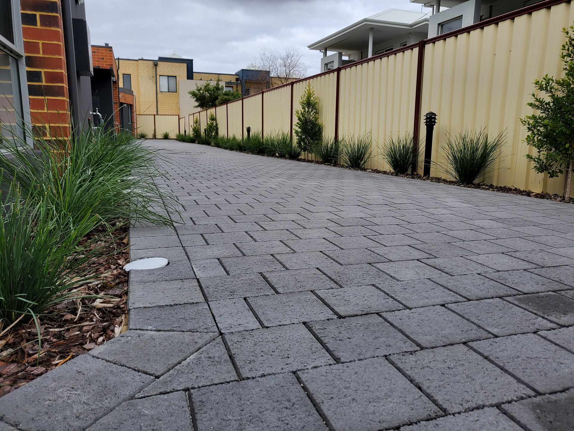 Brick paved walkway with greenery on the left and a beige fence with shrubs on the right.