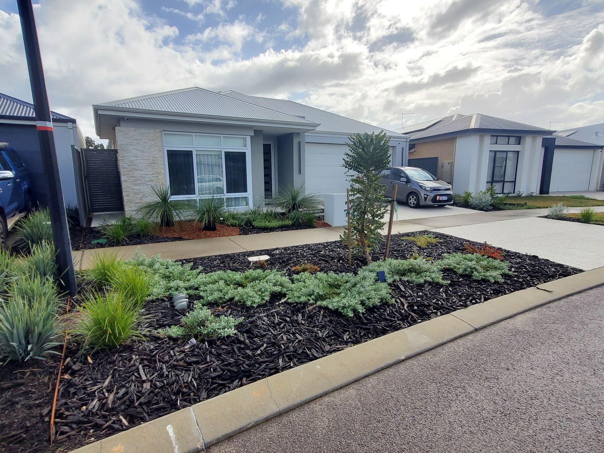 A modern house with a landscaped front yard, featuring dark mulch and various plants, with a cloudy sky.