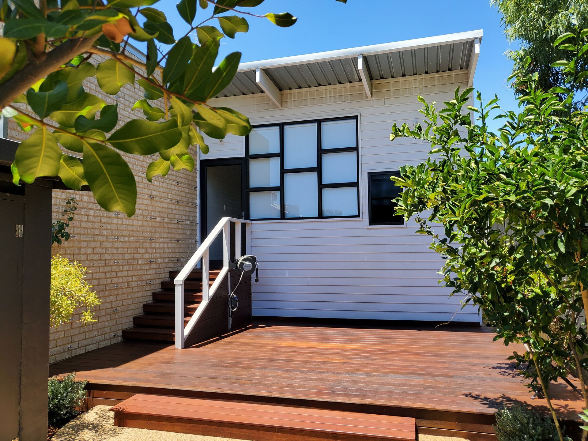 Small house exterior with brick siding, white stairs, and a wooden deck surrounded by greenery