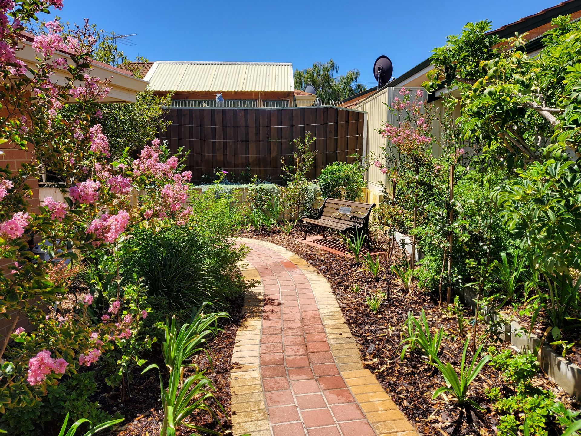 Brick pathway winds through a lush garden with pink flowers, leading toward a wooden fence under a blue sky.
