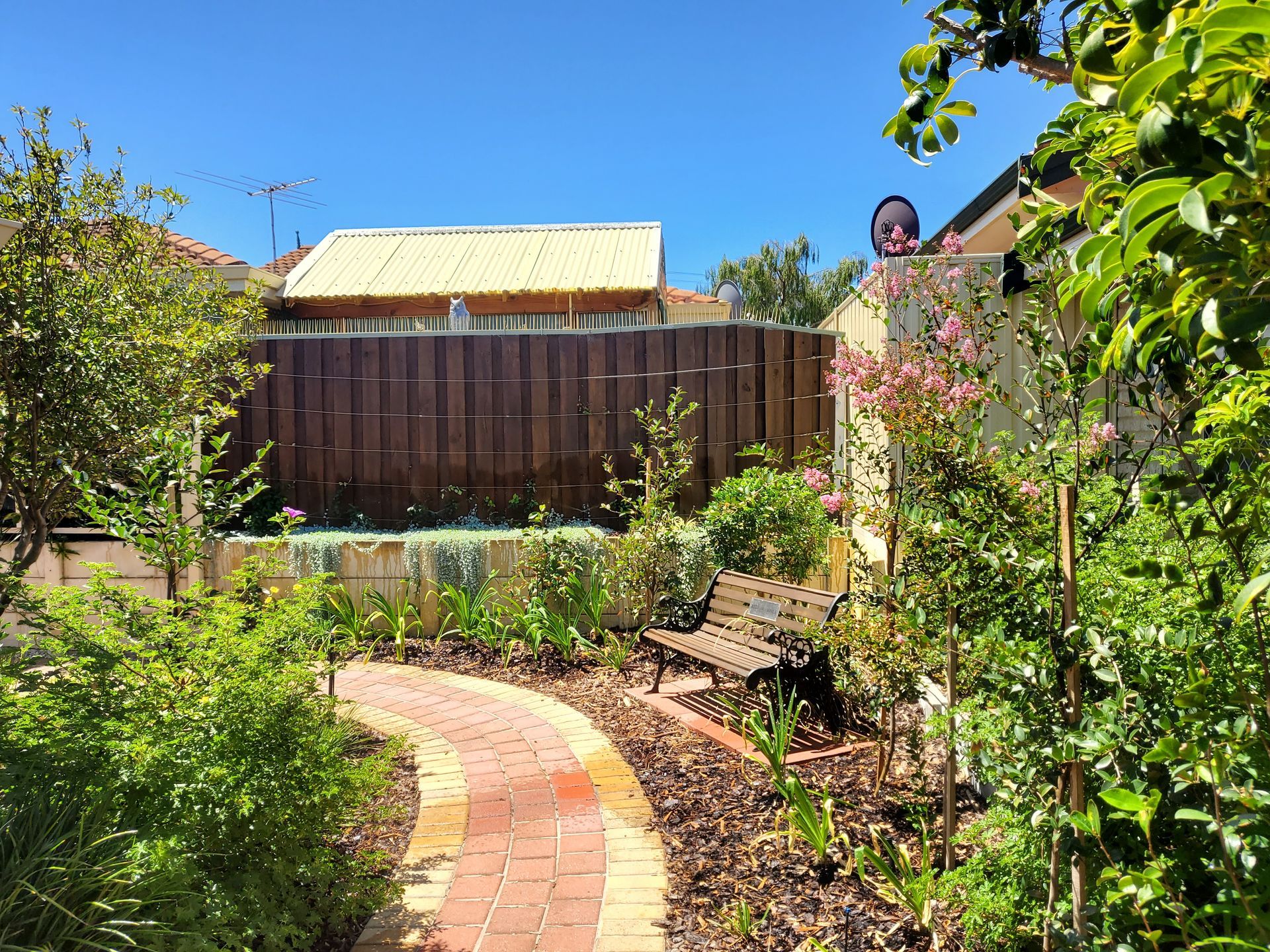 Brick path curves through a lush garden with a wooden bench and fence against a bright blue sky.