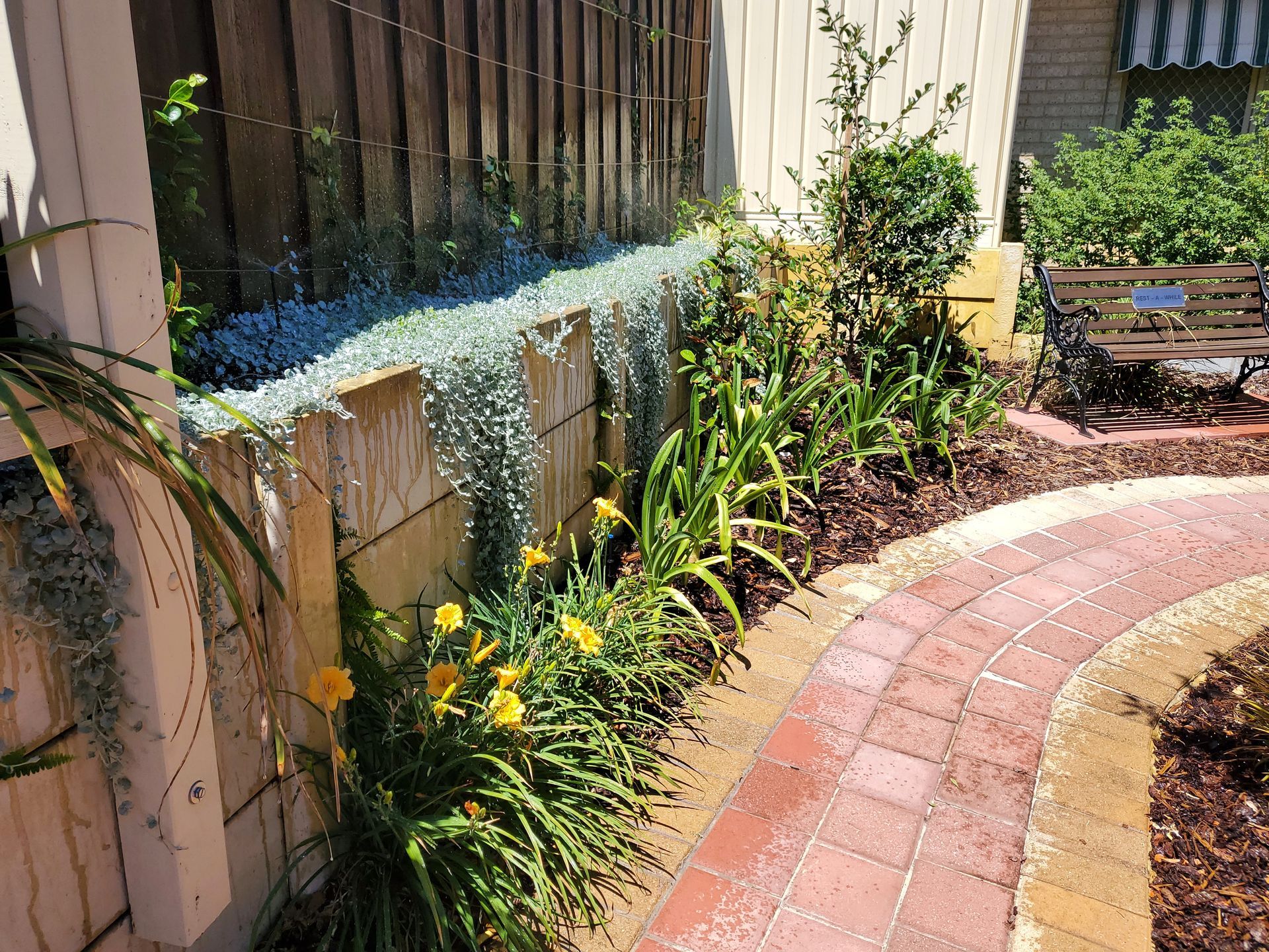 Brick path leads past a garden with flowers and plants next to a wooden fence.