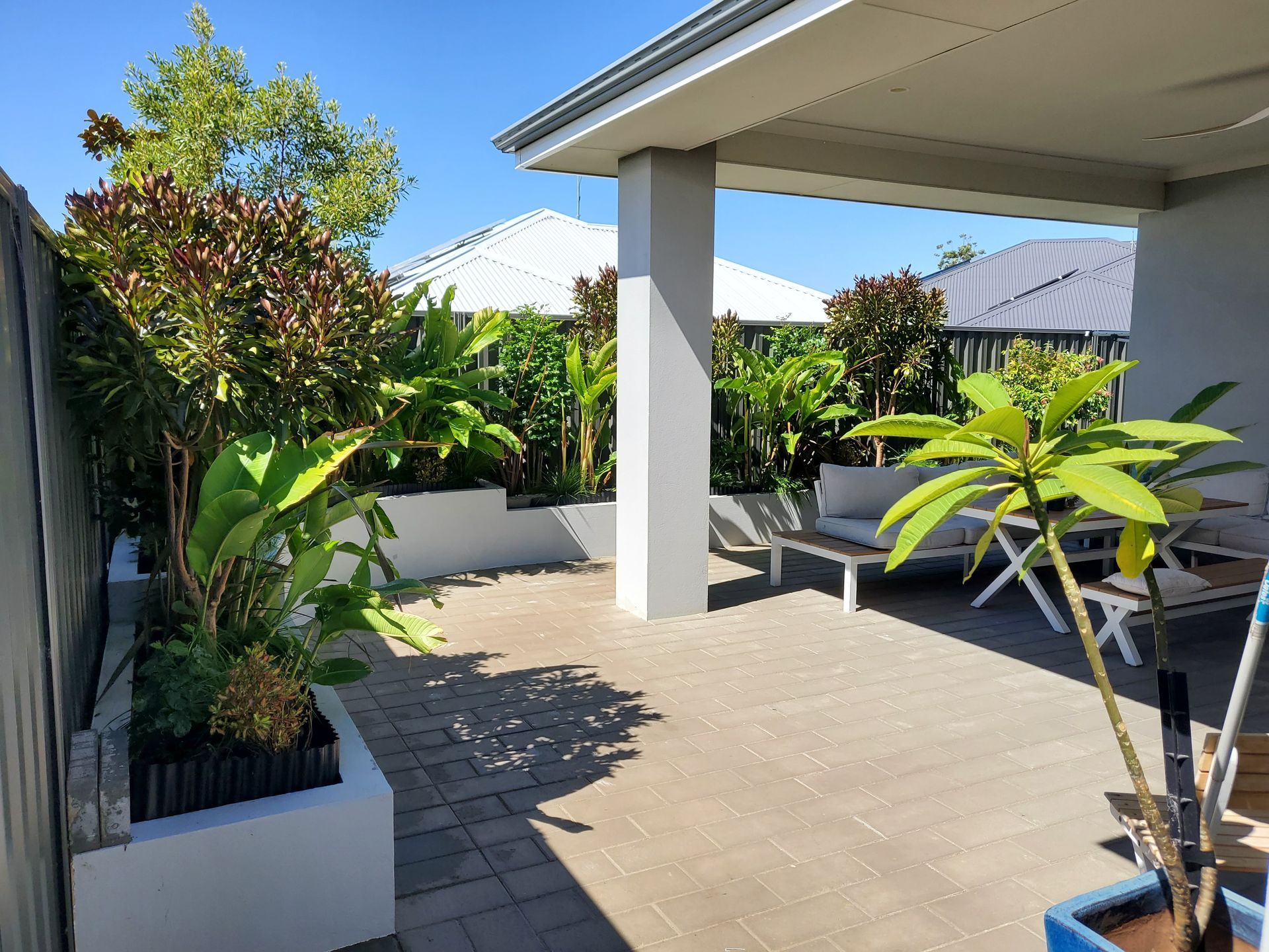A covered patio with a garden and seating area. Green plants and white walls create a bright outdoor space.
