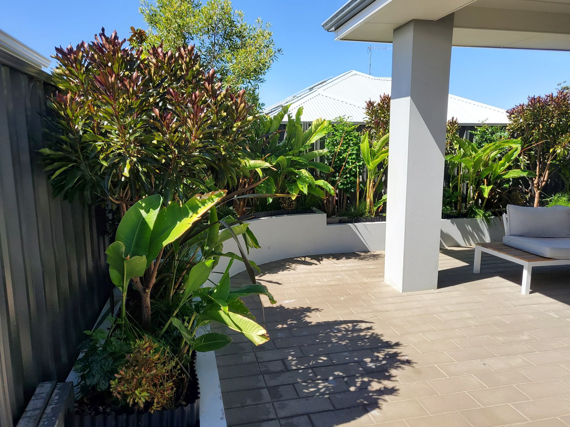A patio with lush greenery and a bench under a white column and roof.