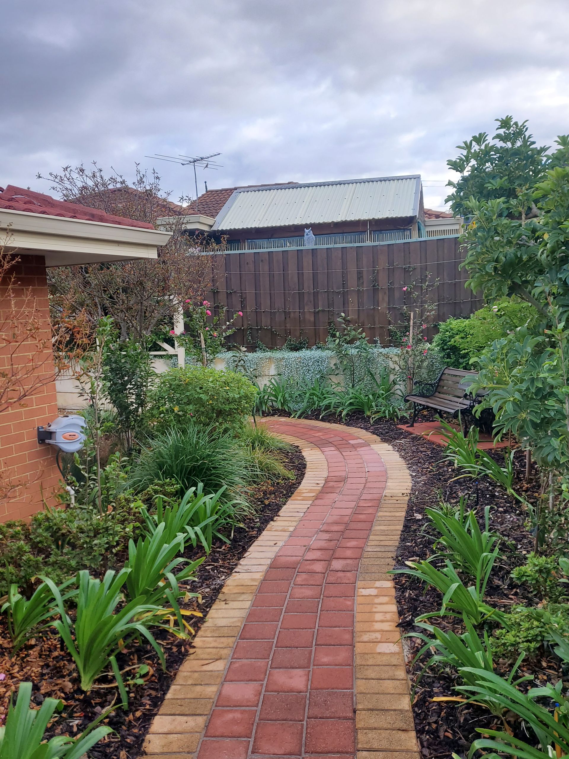 Curved brick garden path beside flower beds and a house, leading to a wooden fence under a cloudy sky