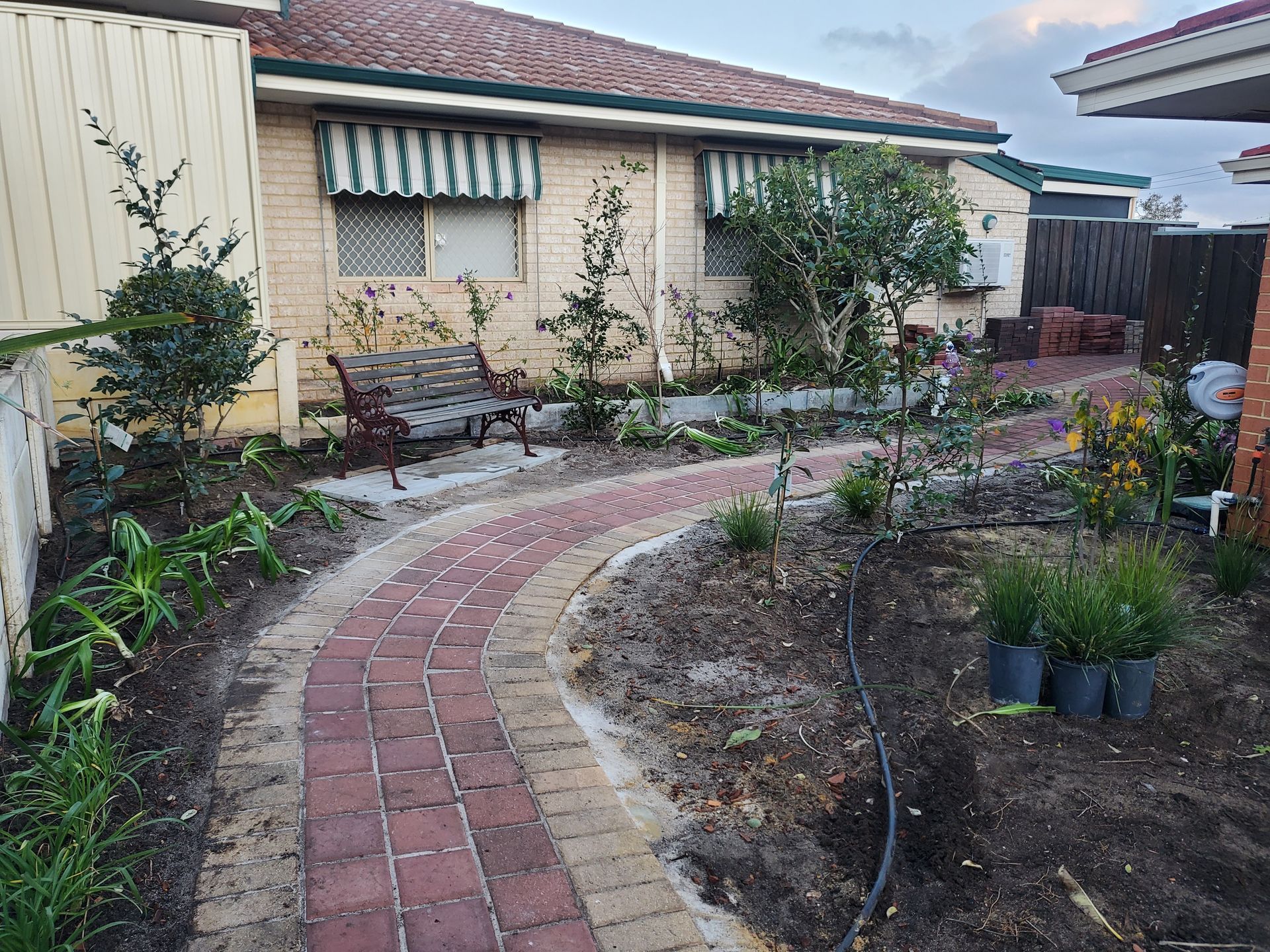 Brick path curves through a garden with a bench, leading to a brick house with green and white awnings.