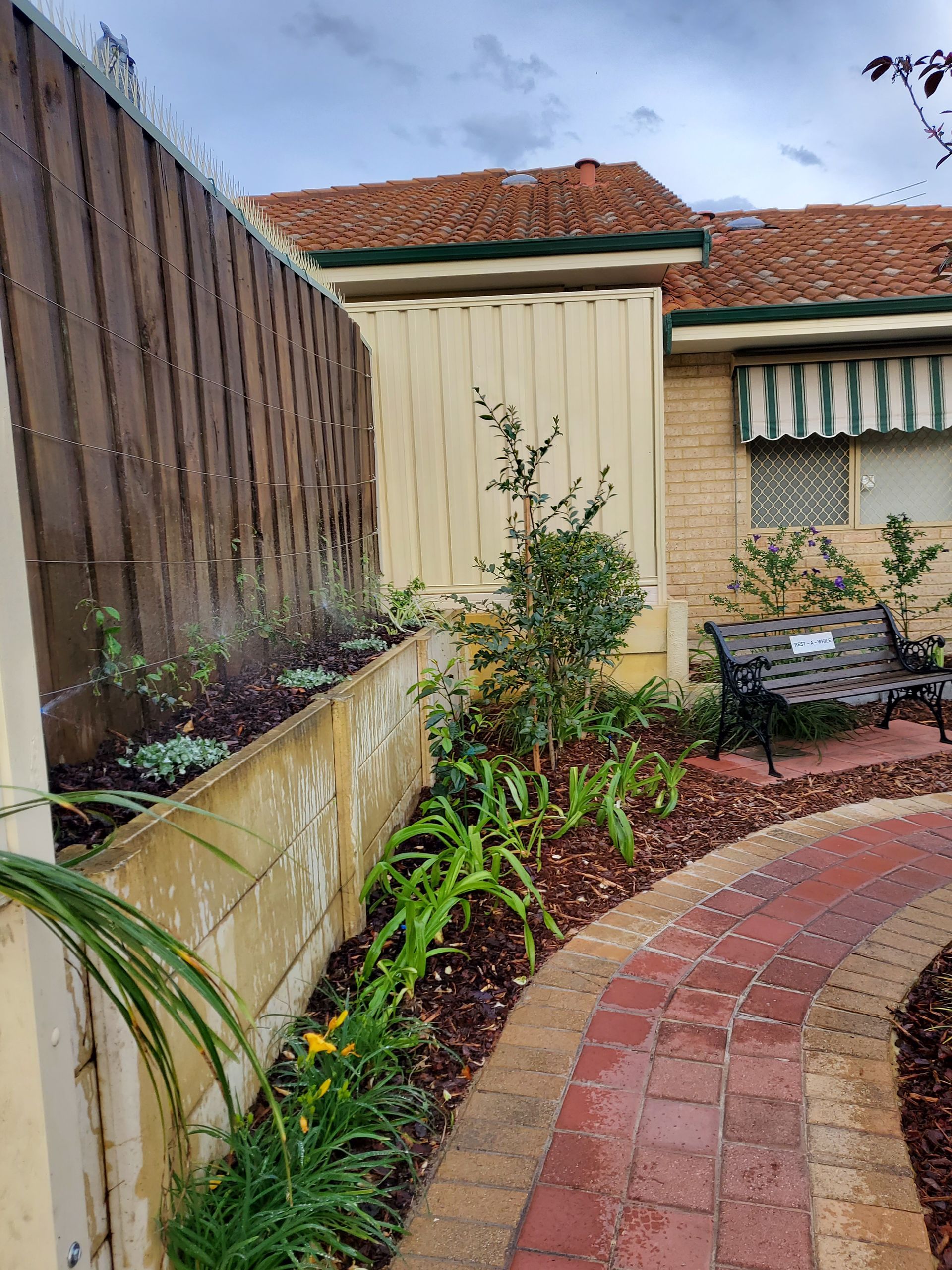 A garden with a brick path, plants, a wooden fence, and a bench against a house.