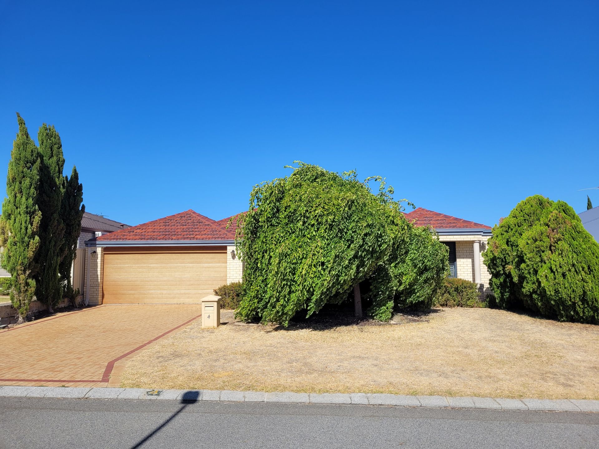 House with tan garage door, brown roof, and front yard with brown gravel and trimmed bushes under a blue sky.