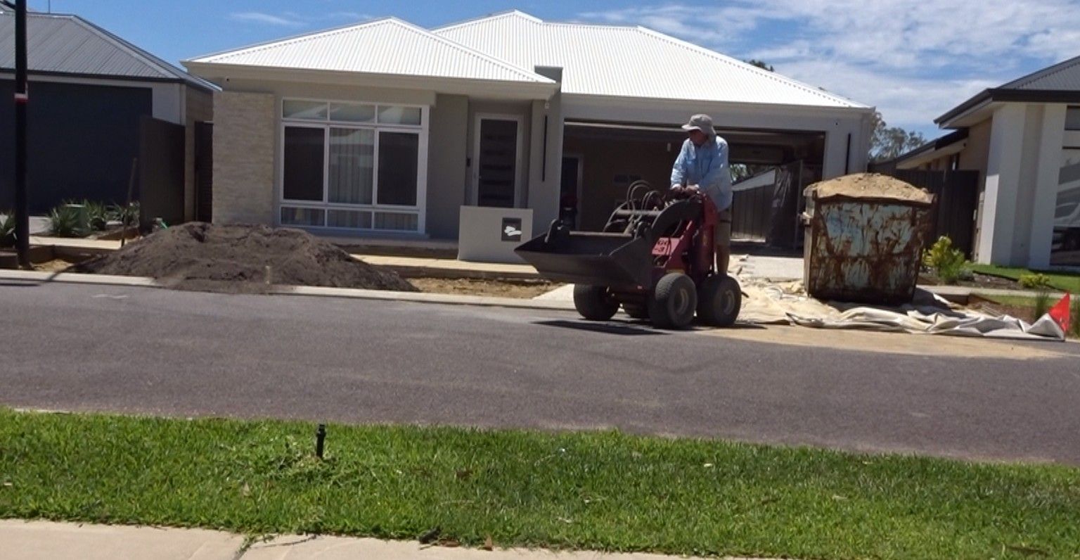 Person operating a small motorized wheelbarrow on a residential street in front of a house under construction.