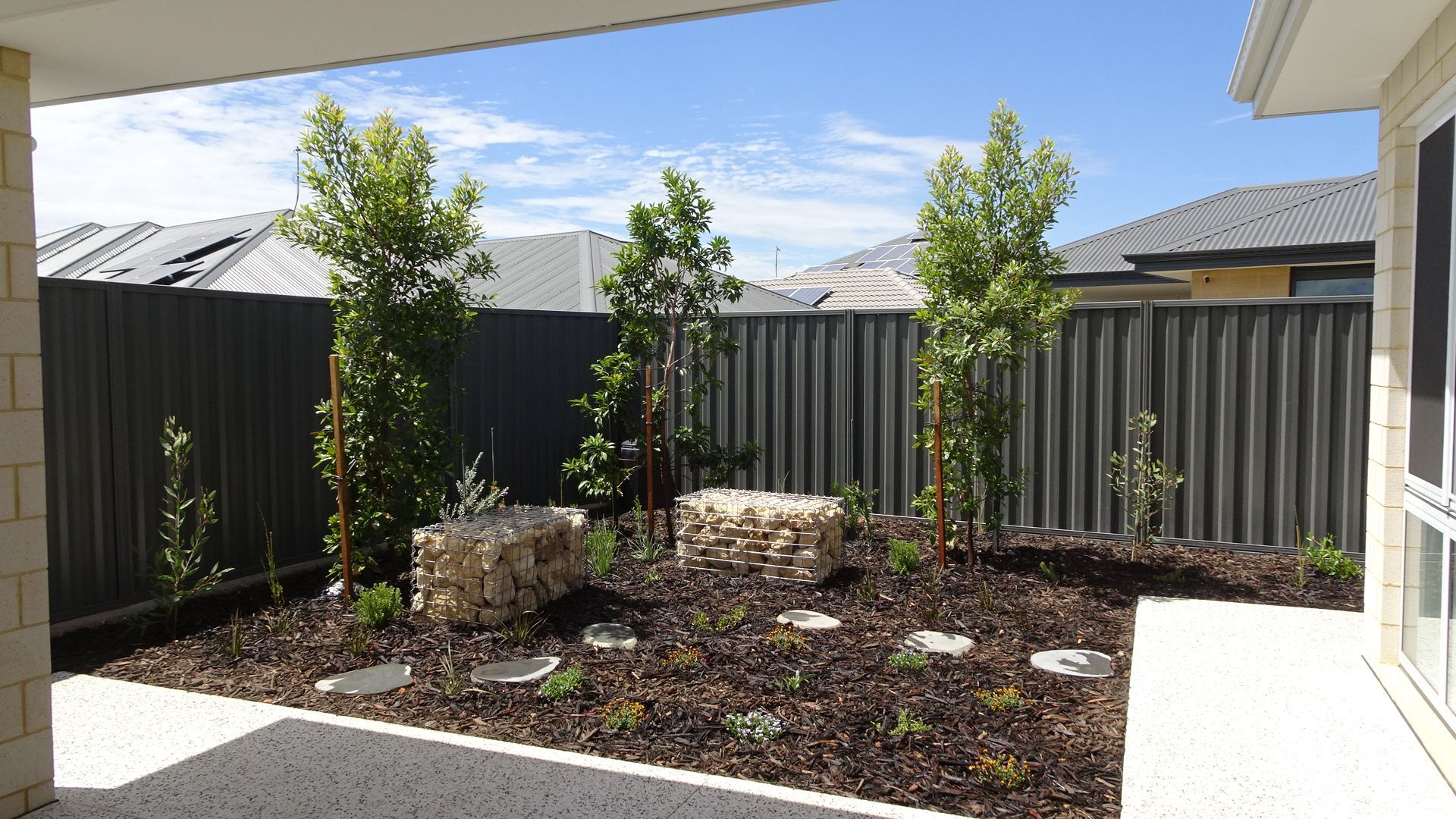Backyard with trees, rocks, and mulch against a dark gray fence, viewed from a covered patio.