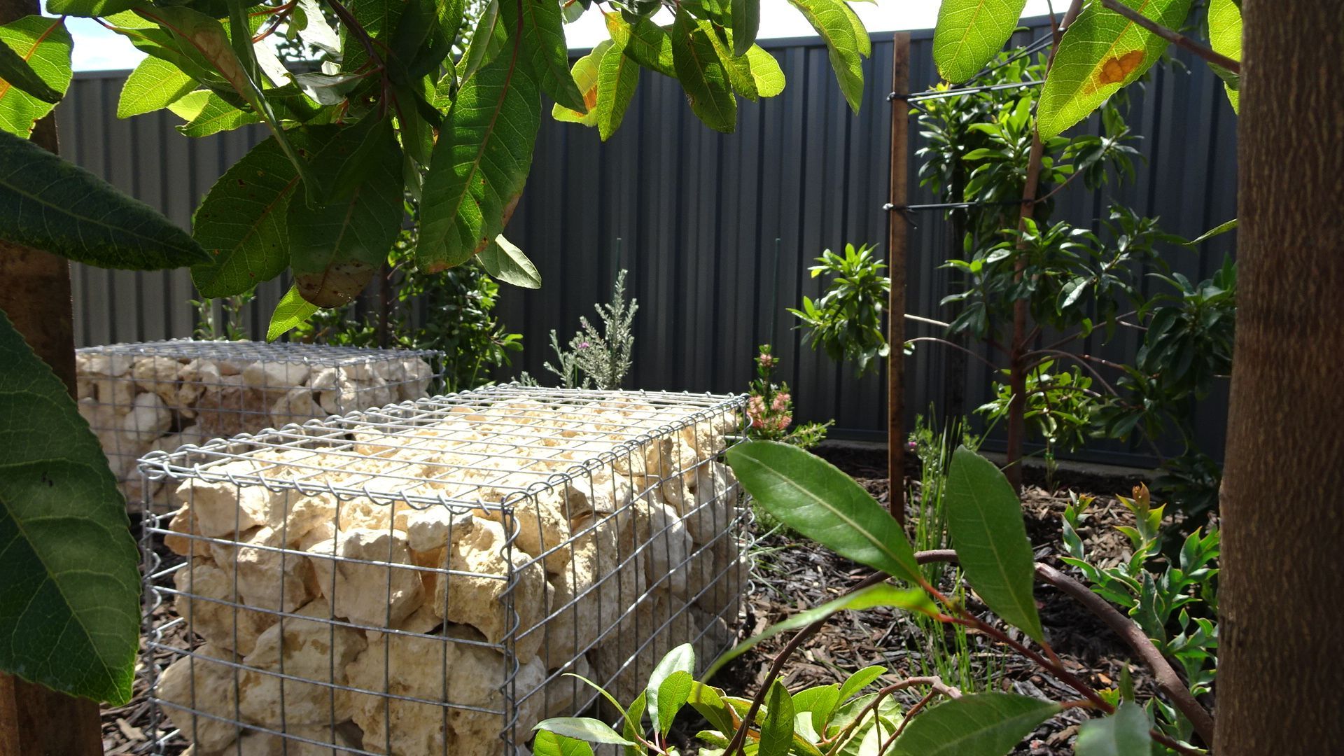 Two stacked stone gabion planters in a garden, framed by green leaves, against a dark fence.