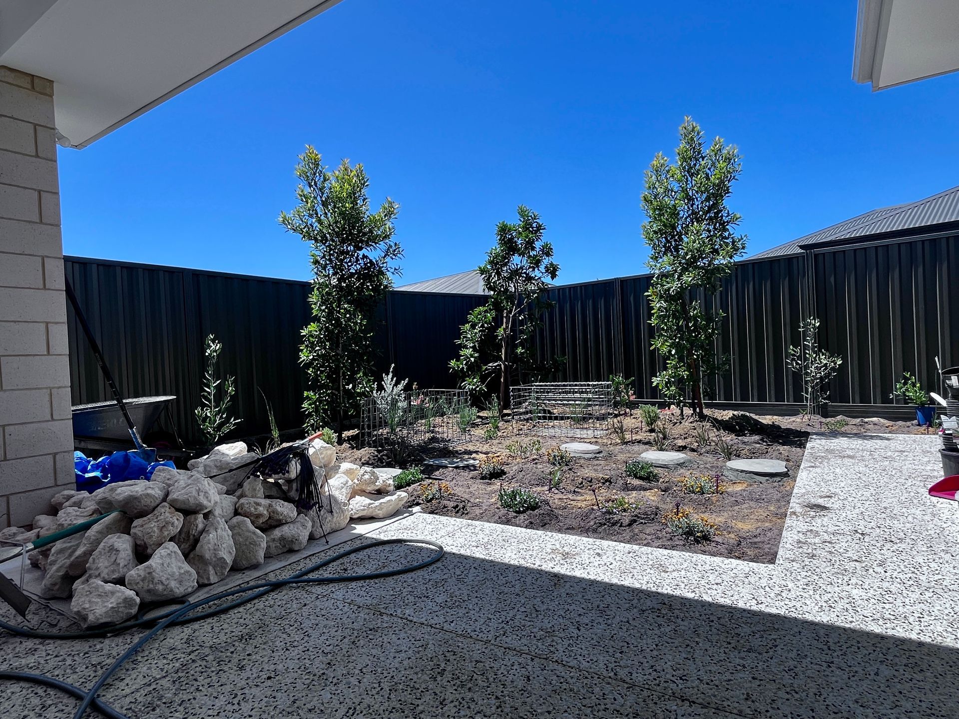Backyard with three trees, gravel patio, dark fence, bright blue sky, and construction debris in foreground.