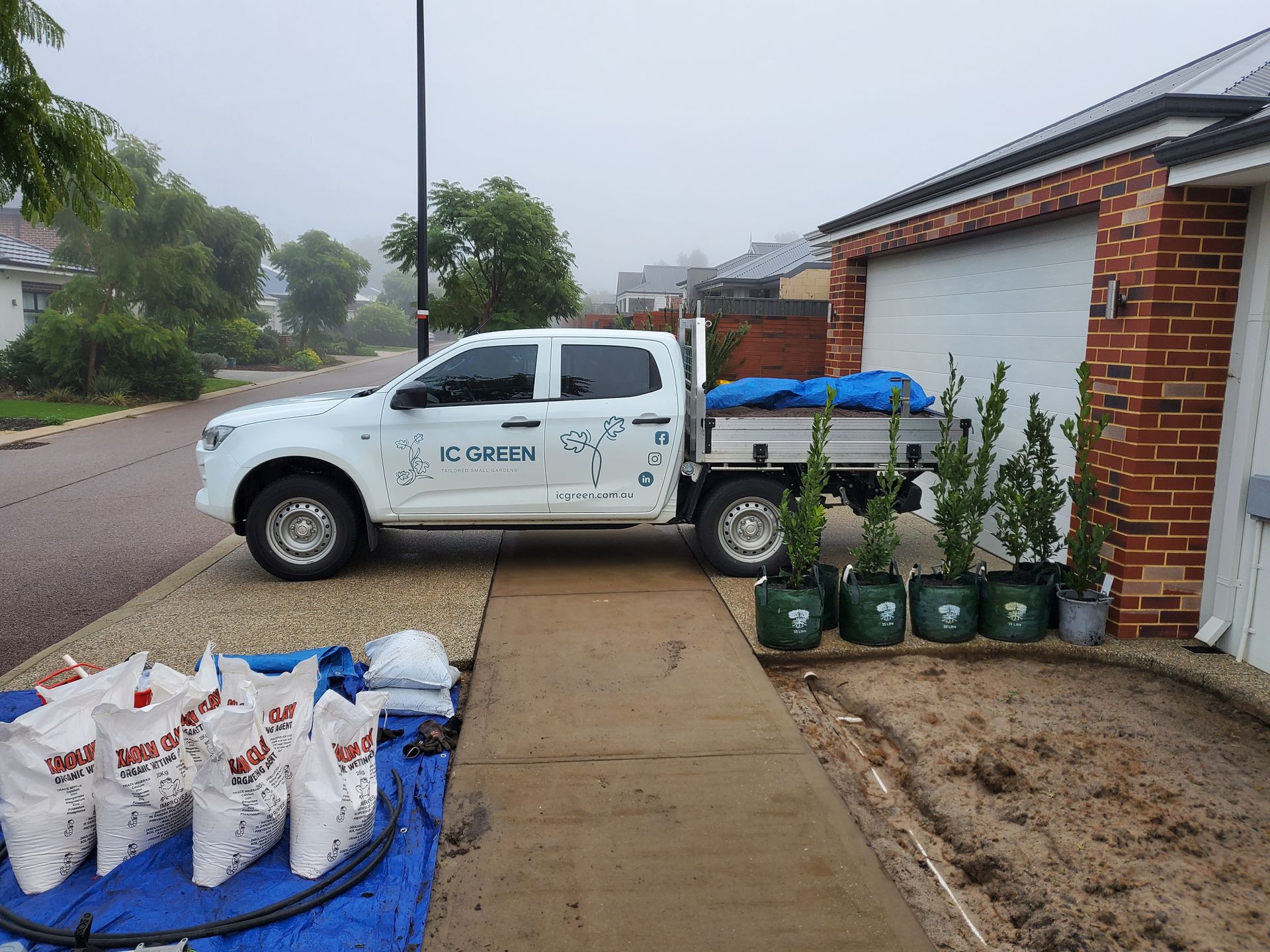 White truck with landscaping supplies parked in front of a house. Bags of soil and potted plants are visible. Cloudy day.