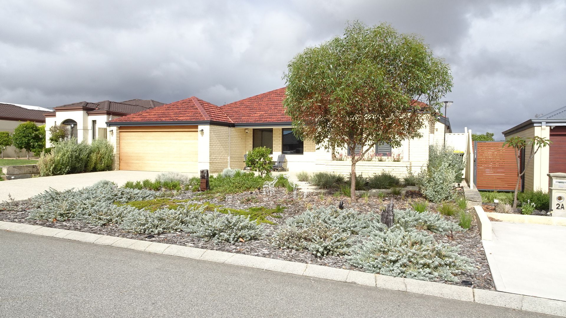 Single-story house with a red tiled roof, cream-colored walls, and a front yard with small plants and a tree; cloudy sky.