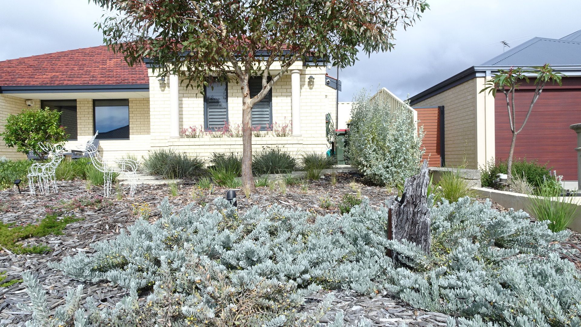 Residential landscape featuring a house with a red roof, various plants, and a sign.