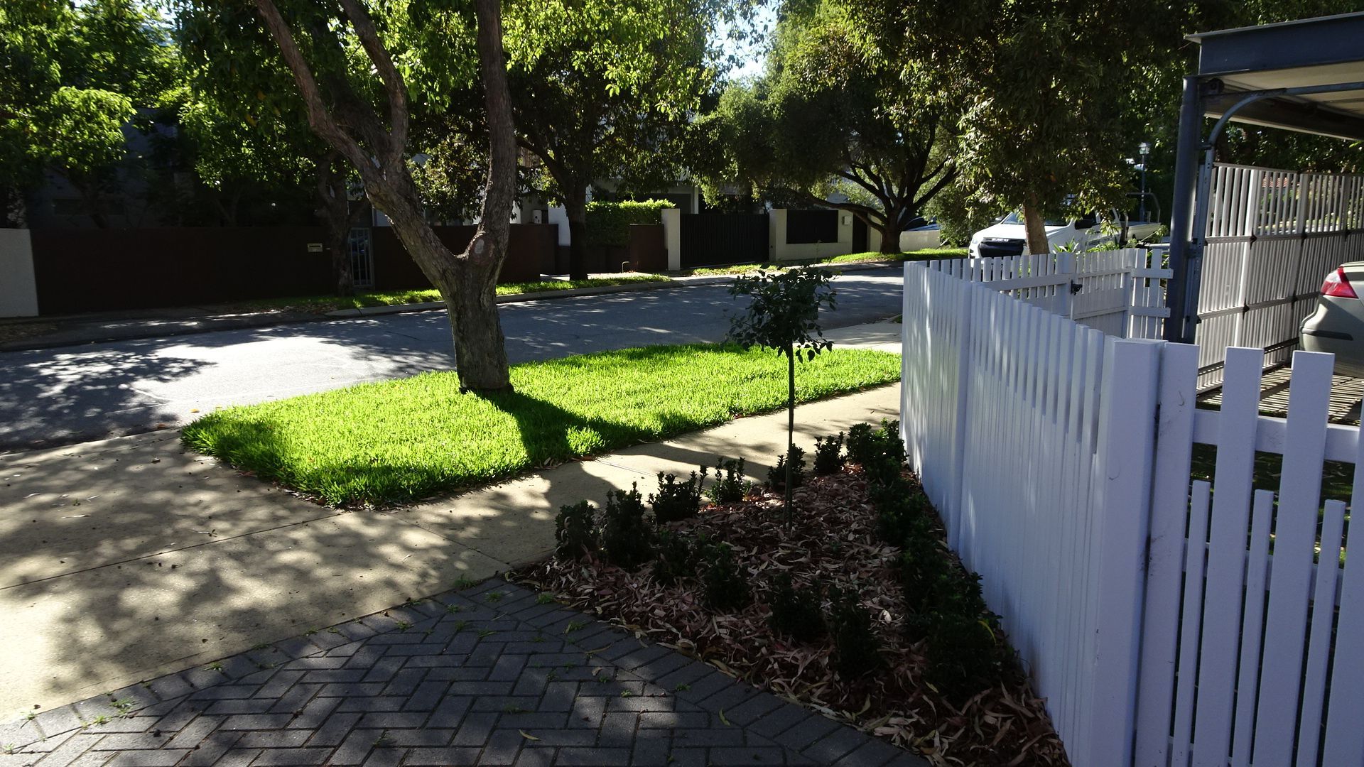 Street scene with green grass, trees, white picket fence, and a brick driveway. Sunny day.