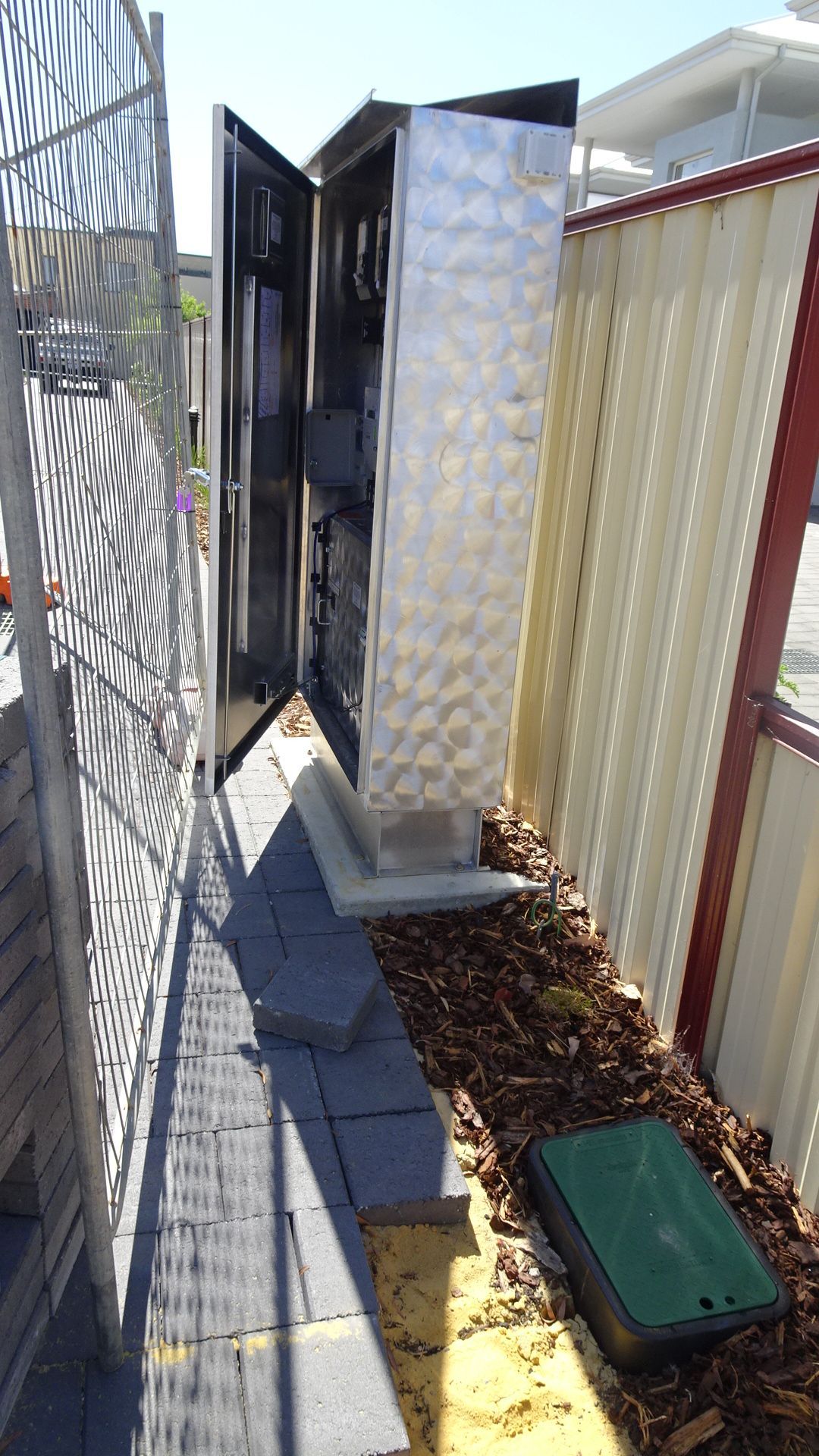 Metal electrical box with open doors, between a fence and a walkway.