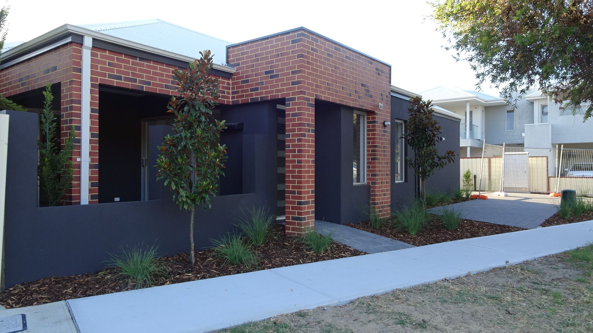 Modern one-story house with red brick accents and dark grey exterior. Sidewalk and landscaping in front.