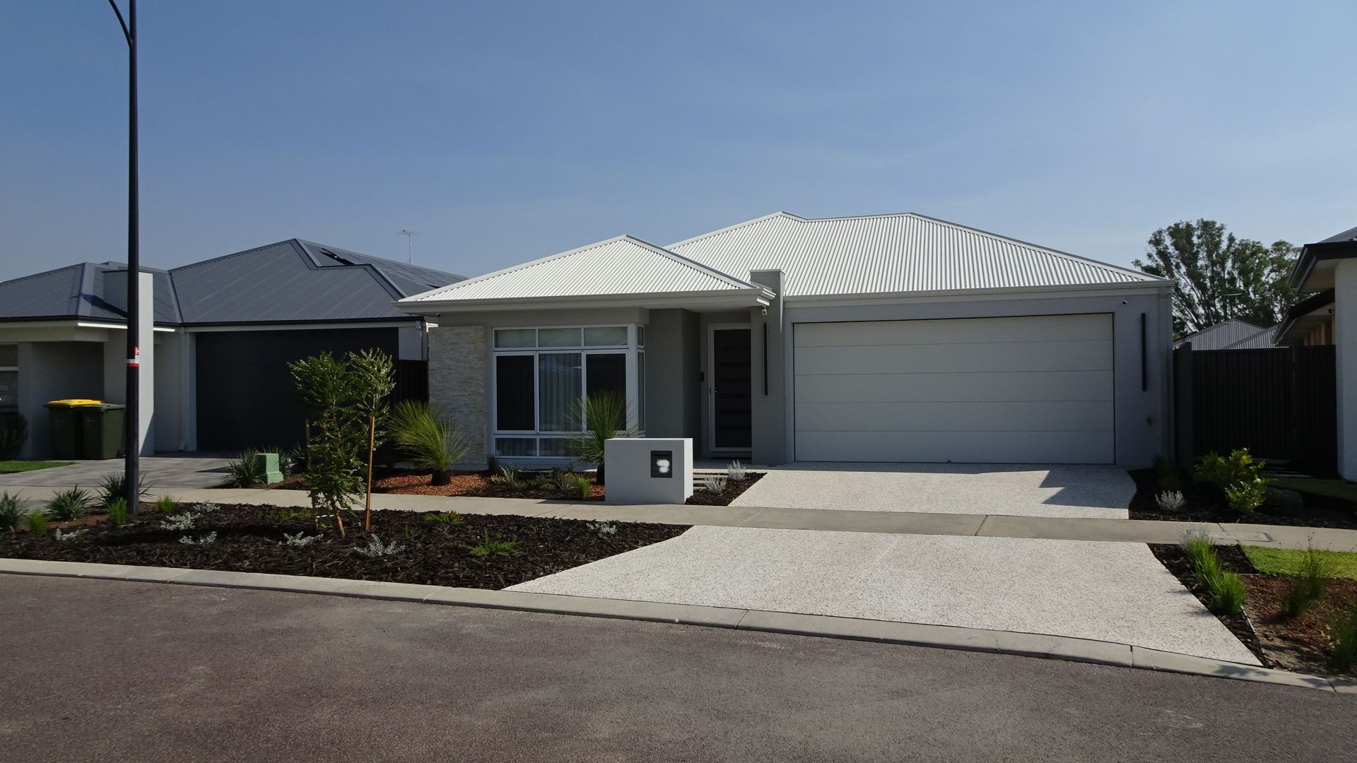 Modern single-story house with light gray facade, white roof, and gravel driveway. Blue sky.