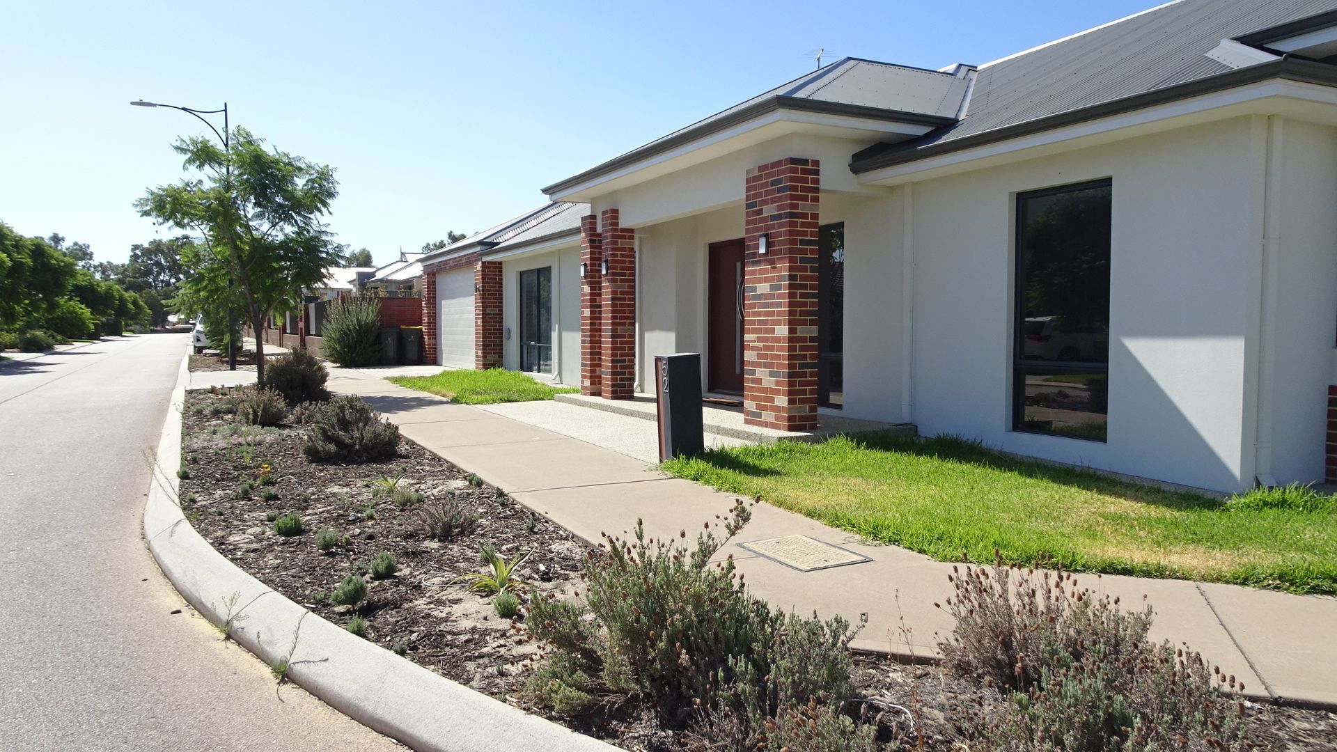 A row of white houses with brick columns and dark windows, a sidewalk with landscaping, and a street.