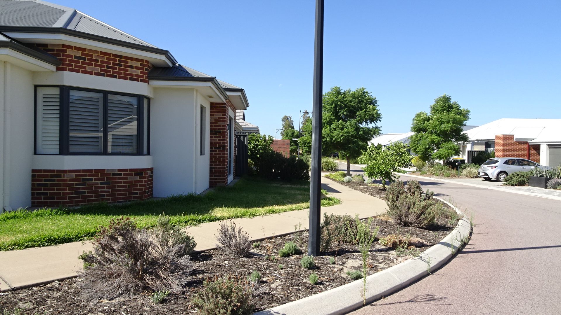Suburban street with houses, a paved walkway, and a car parked in the distance under a blue sky.