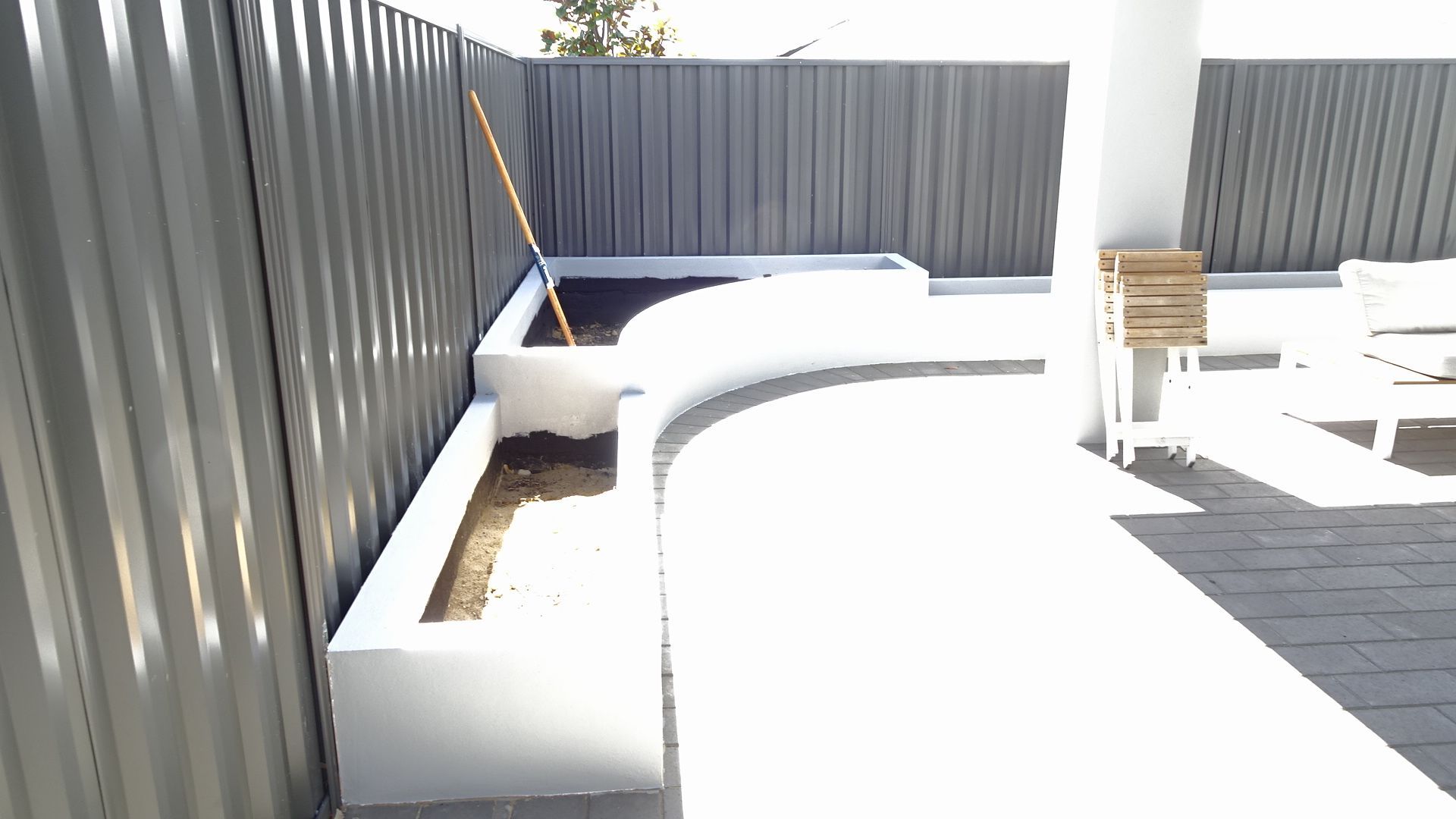 White curved planters alongside a corrugated gray fence. A bright patio is in the sun.