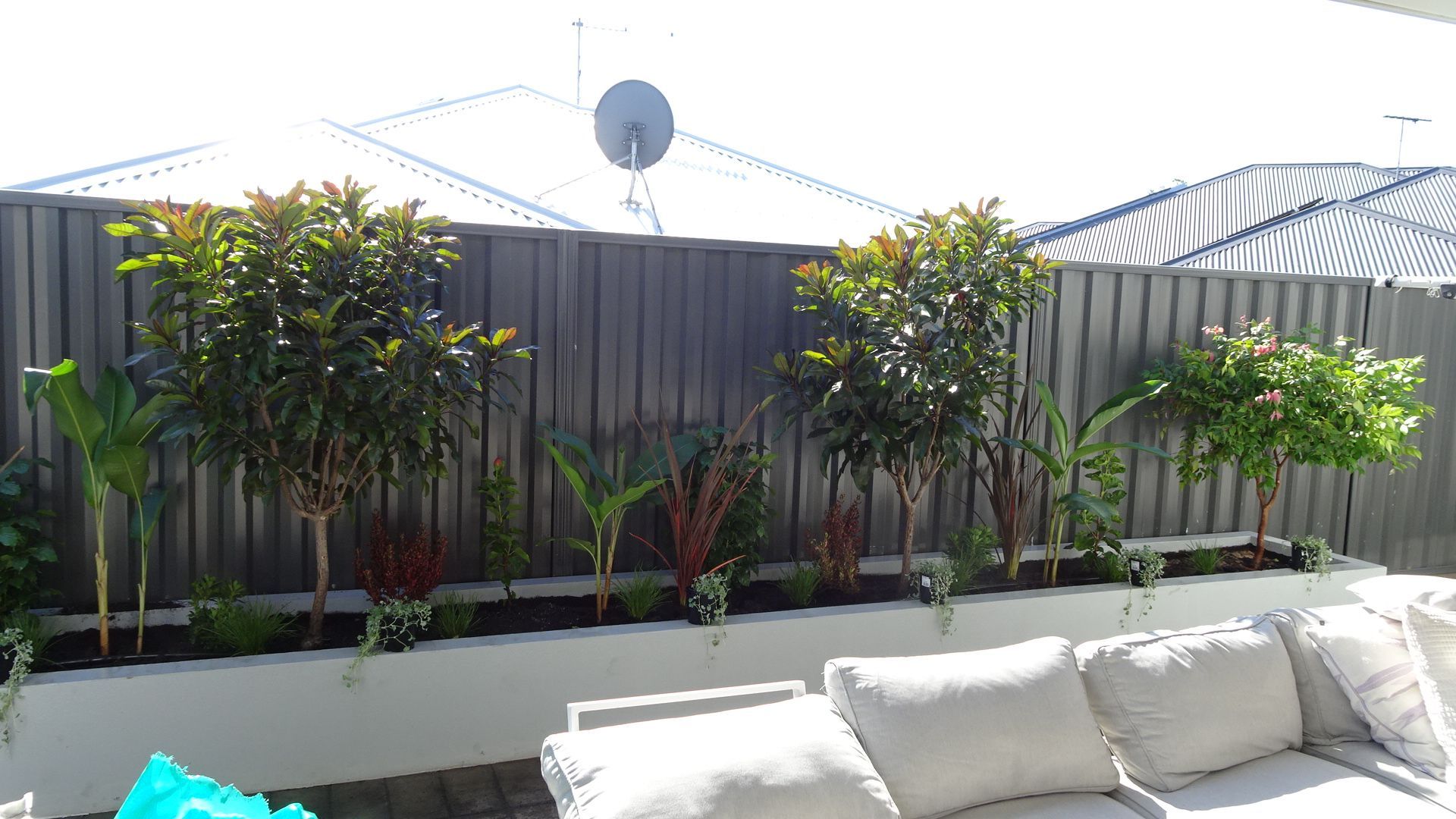 A well-landscaped backyard with a raised planter bed against a gray fence, trees, and a white outdoor sectional sofa.