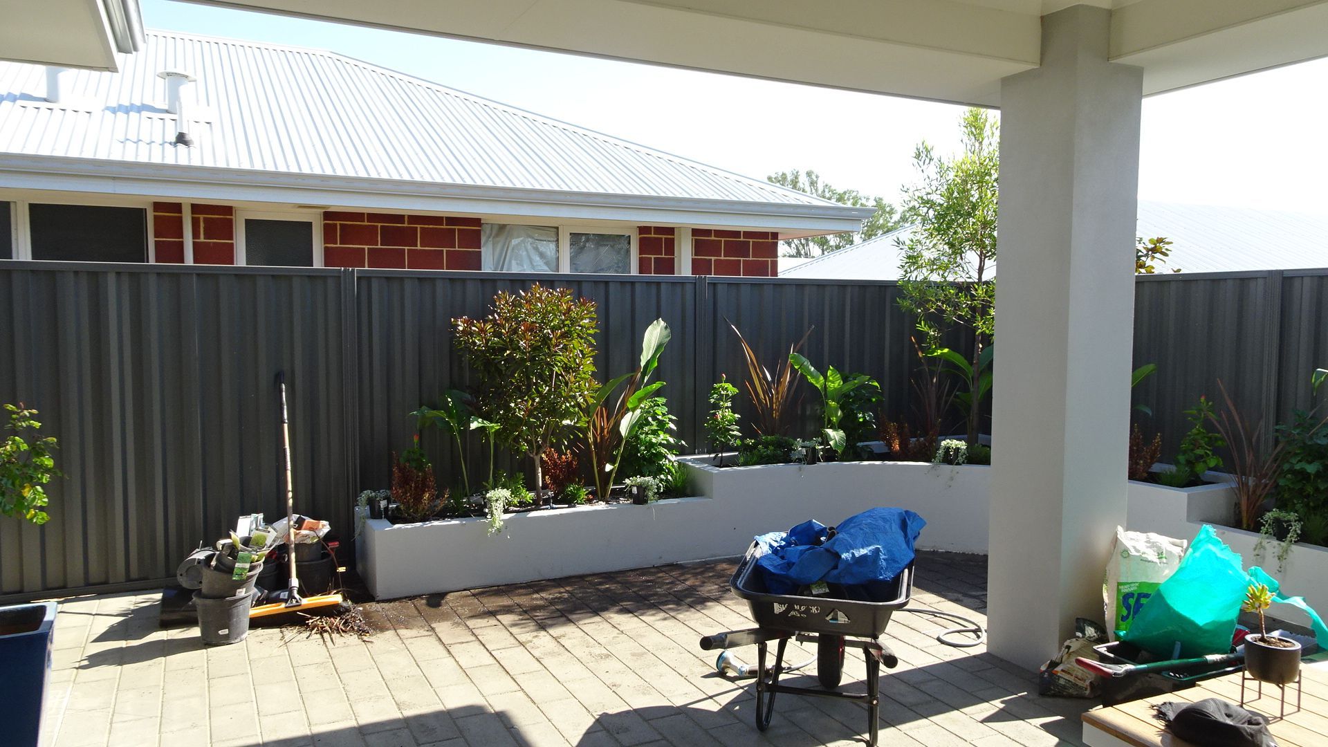 Backyard patio with a garden bed along a gray fence. Brick pavers, a grill, and plants.