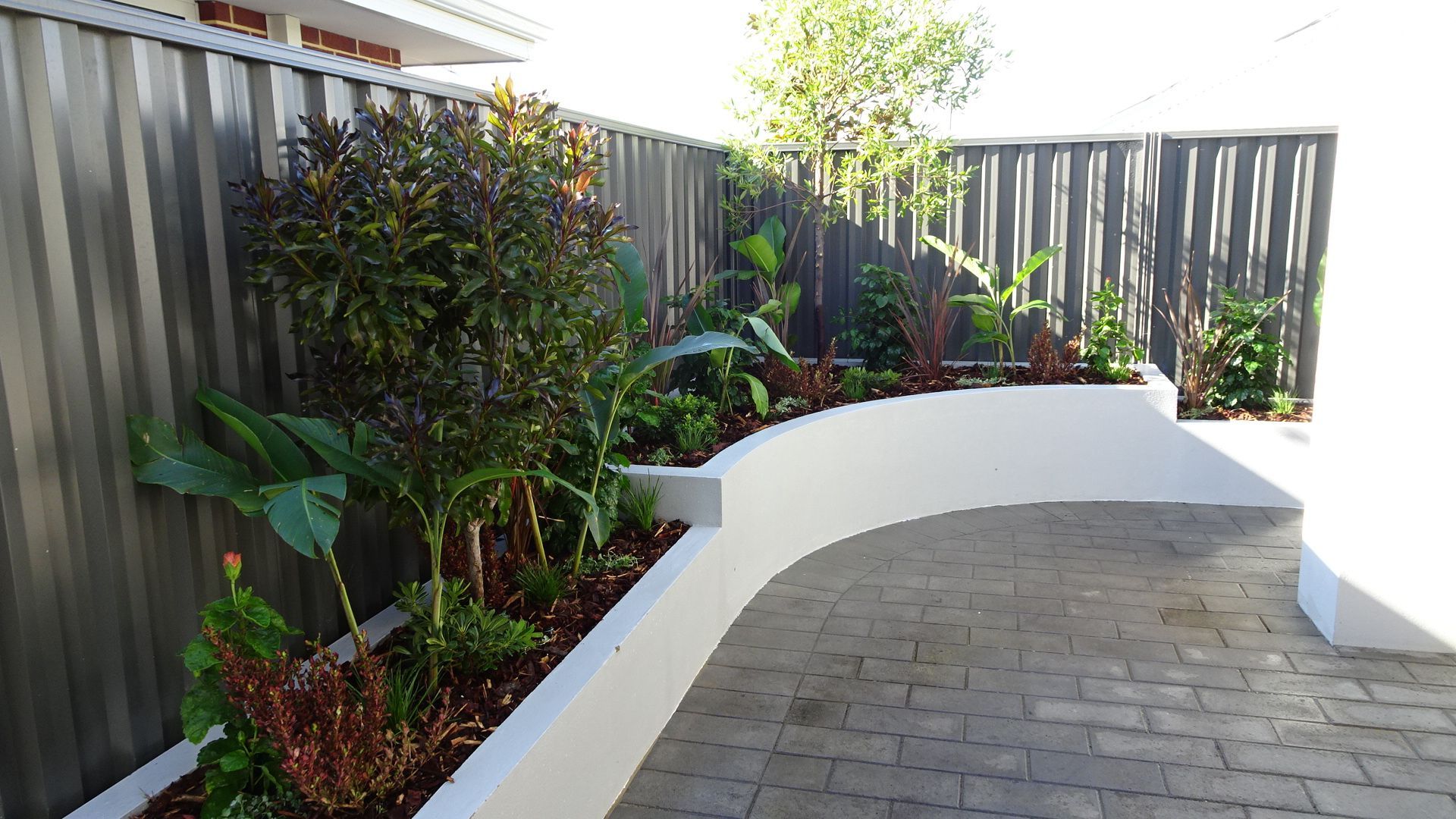 Raised garden bed with lush greenery, against a dark fence and paved pathway.