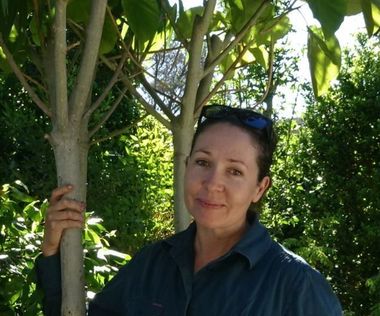 Woman in green shirt, smiling, standing by tree in a sunny garden.