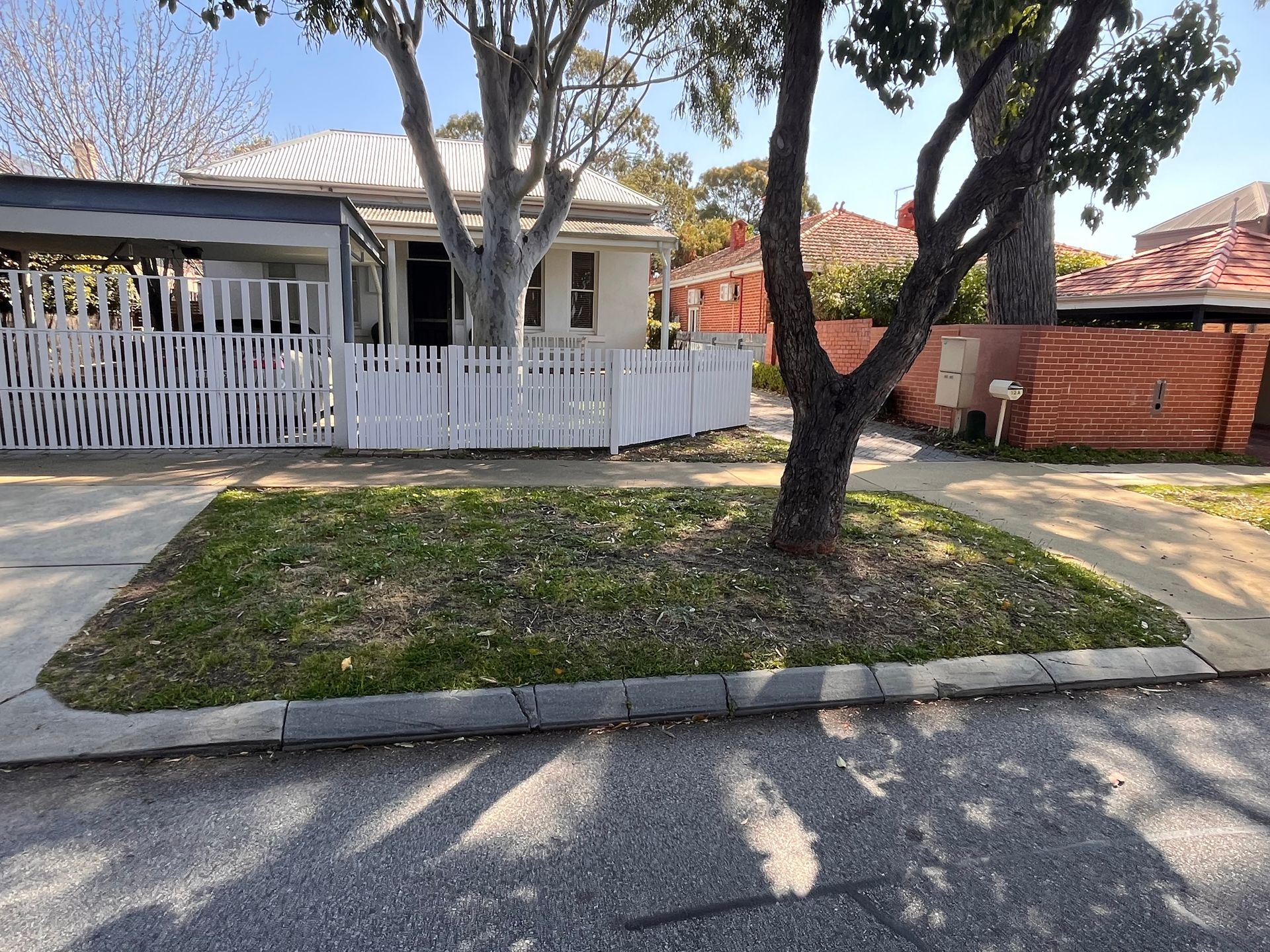 A small house with white picket fence and tree on a grassy patch along a sunny street.