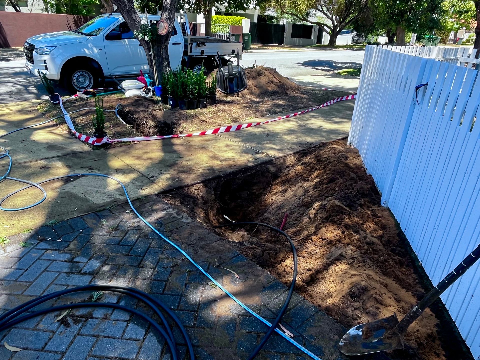 A white truck near a dig site, fenced by a white picket fence. Groundwork with hoses and red/white tape.