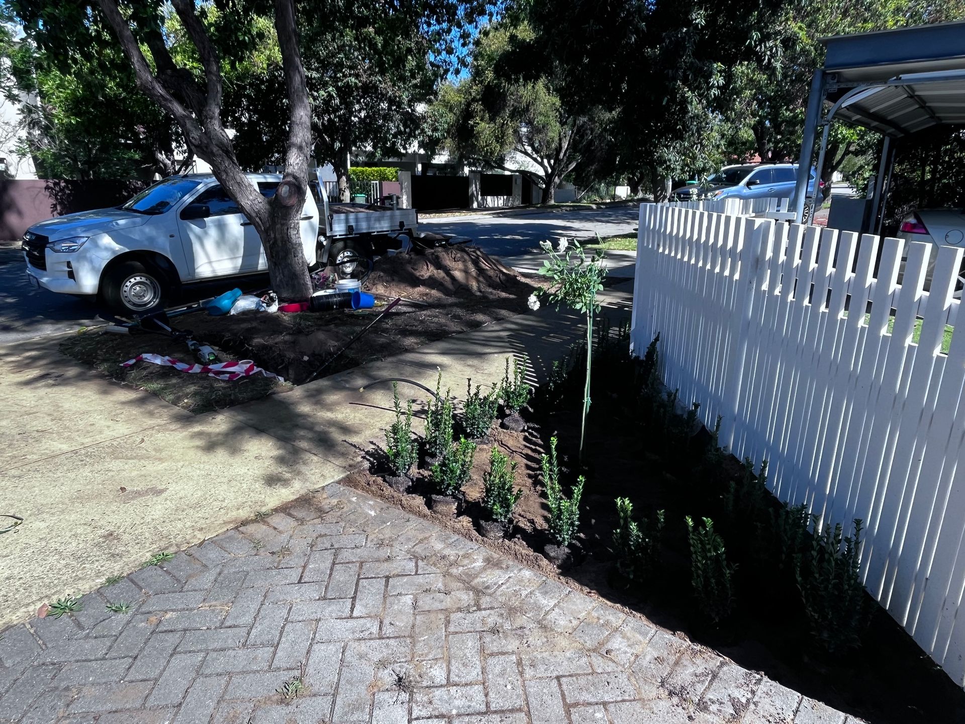 White pickup truck parked near a tree and garden bed next to a white picket fence on a sunny street.