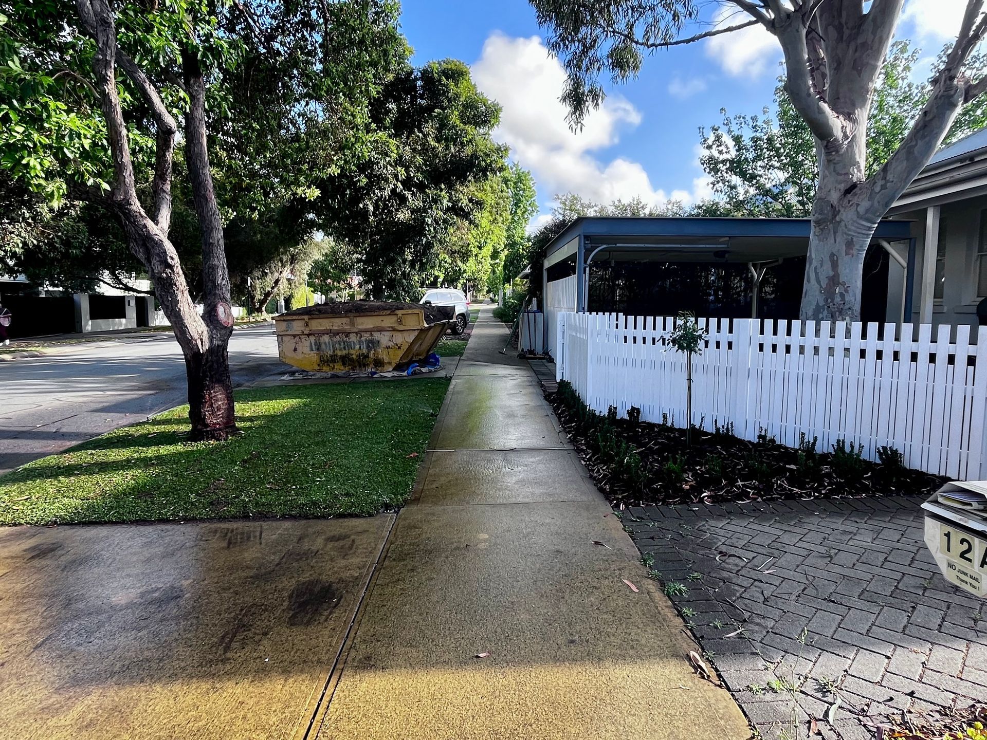 Sidewalk with wet pavement. Trees and green grass, a white picket fence and yellow skip bin are present. Blue sky.