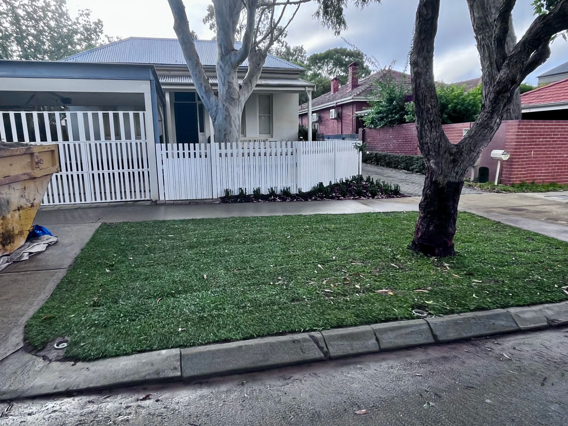 Green lawn in front of a white picket fence, house in background. Tree on the right.