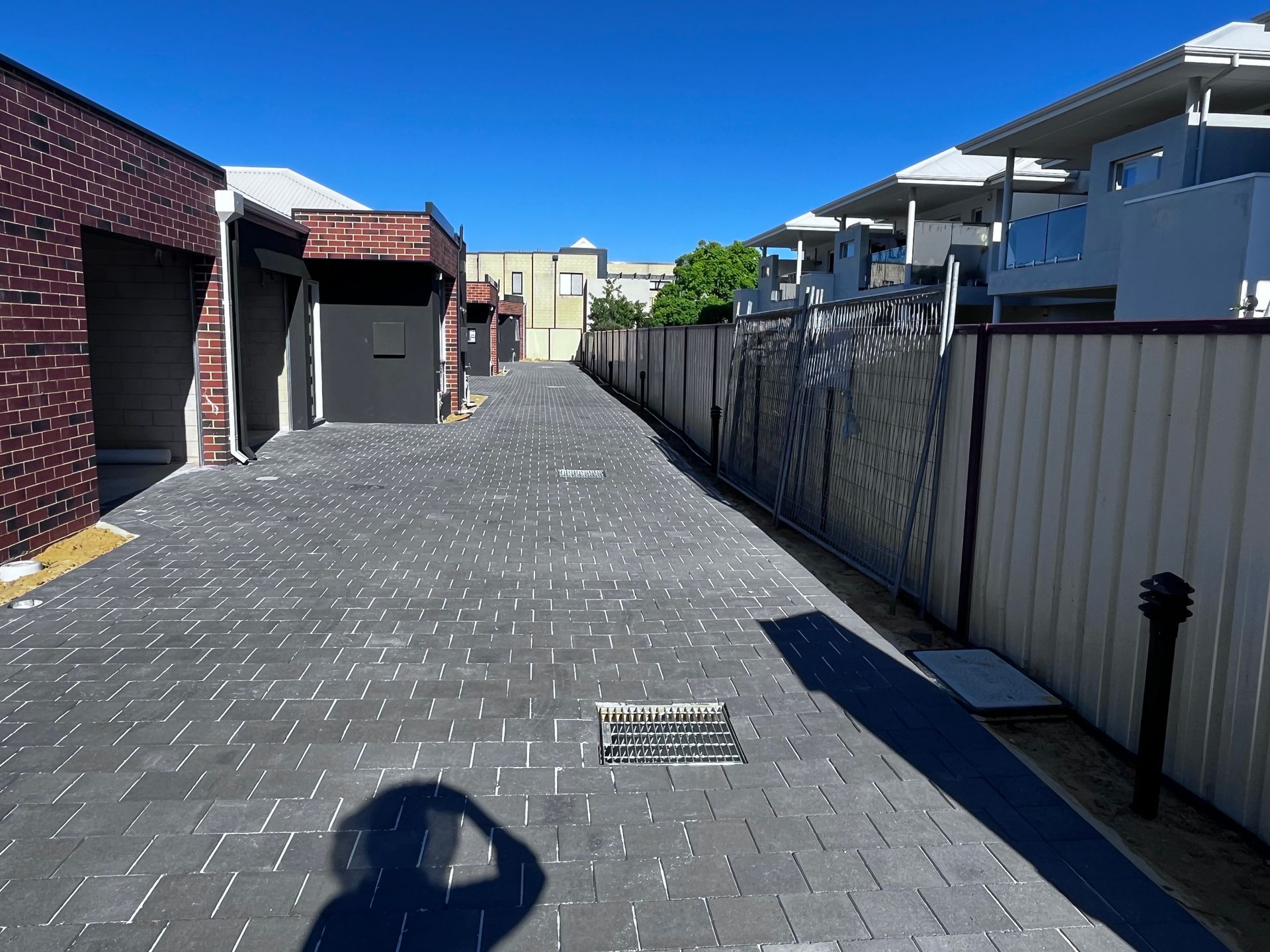 Brick paved alley with buildings and fences under a bright blue sky. Shadow of person in foreground.