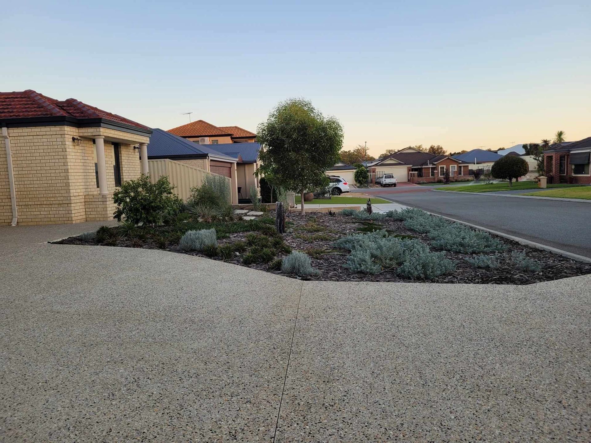 Residential street scene with houses, a garden bed, and a gravel driveway.