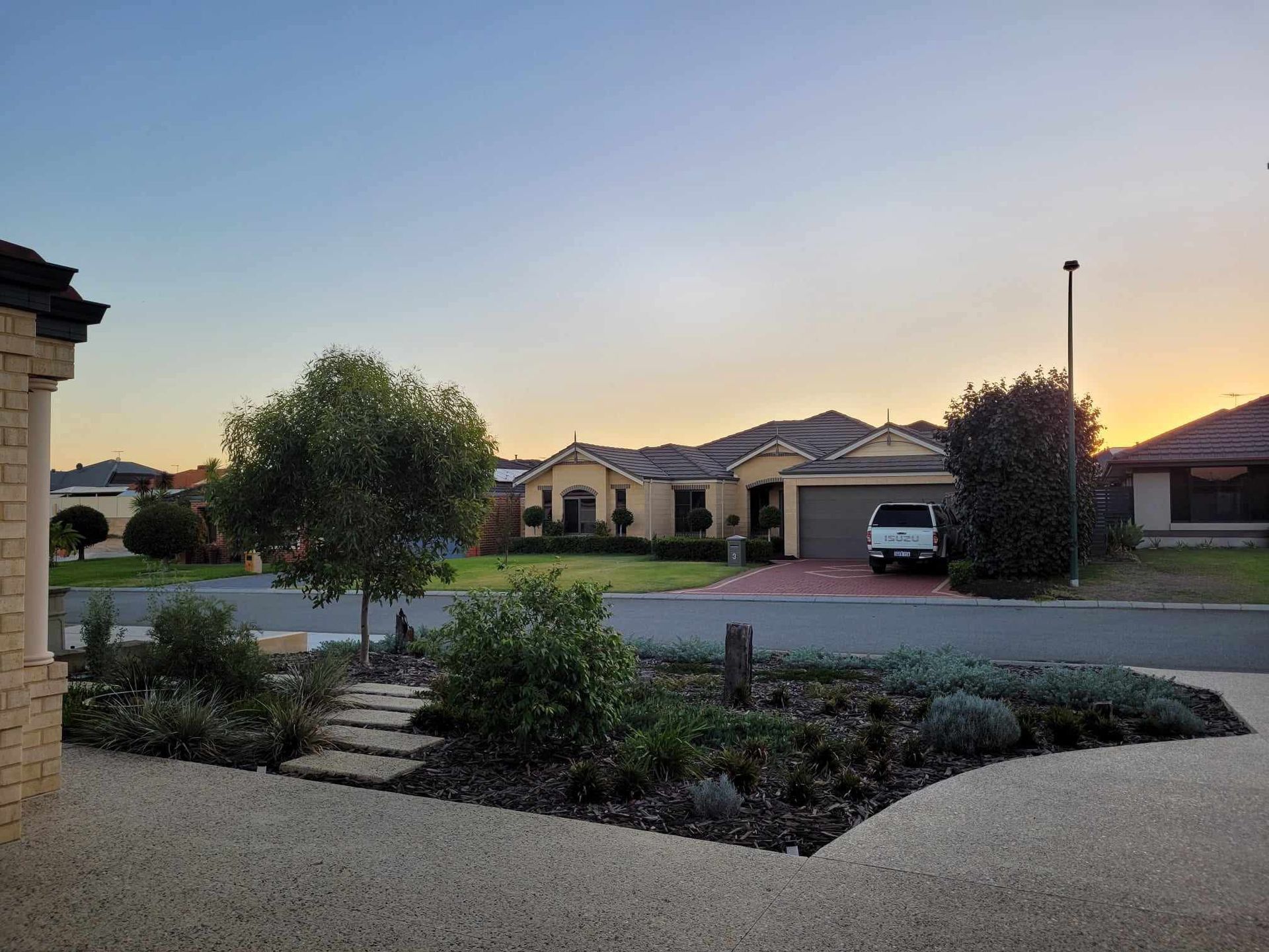 Suburban homes at dusk with a tree-lined street and front yard landscaping; a white truck is parked in a driveway.